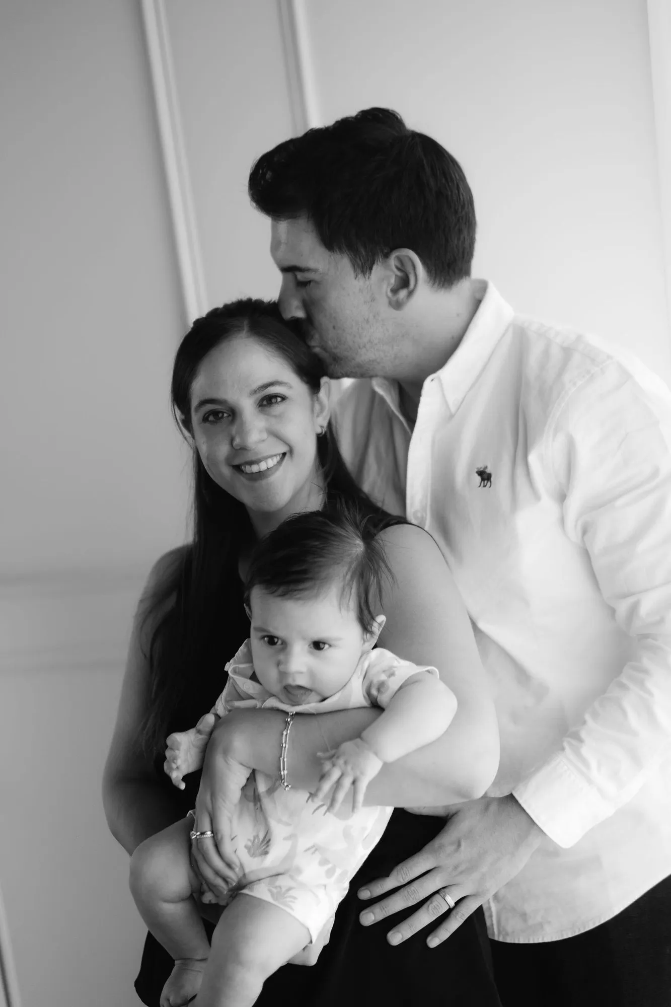 En blanco y negro, un papá besa la cabeza de la mamá, que sostiene a su bebé.