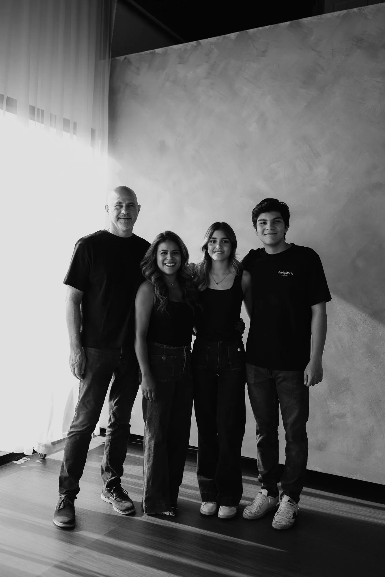 En blanco y negro, una familia de cuatro posa de pie en el estudio, frente a una pared texturizada.