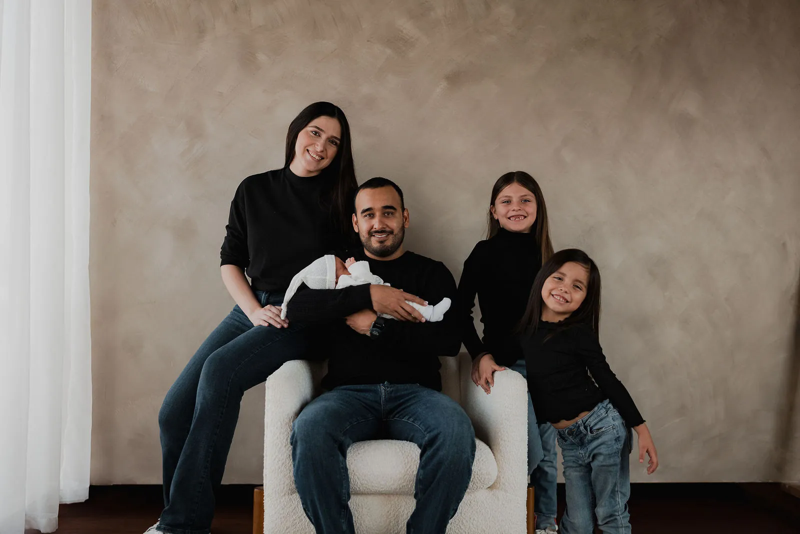 Una familia con un bebé posa sentada en un sillón blanco, frente a una pared color arena.