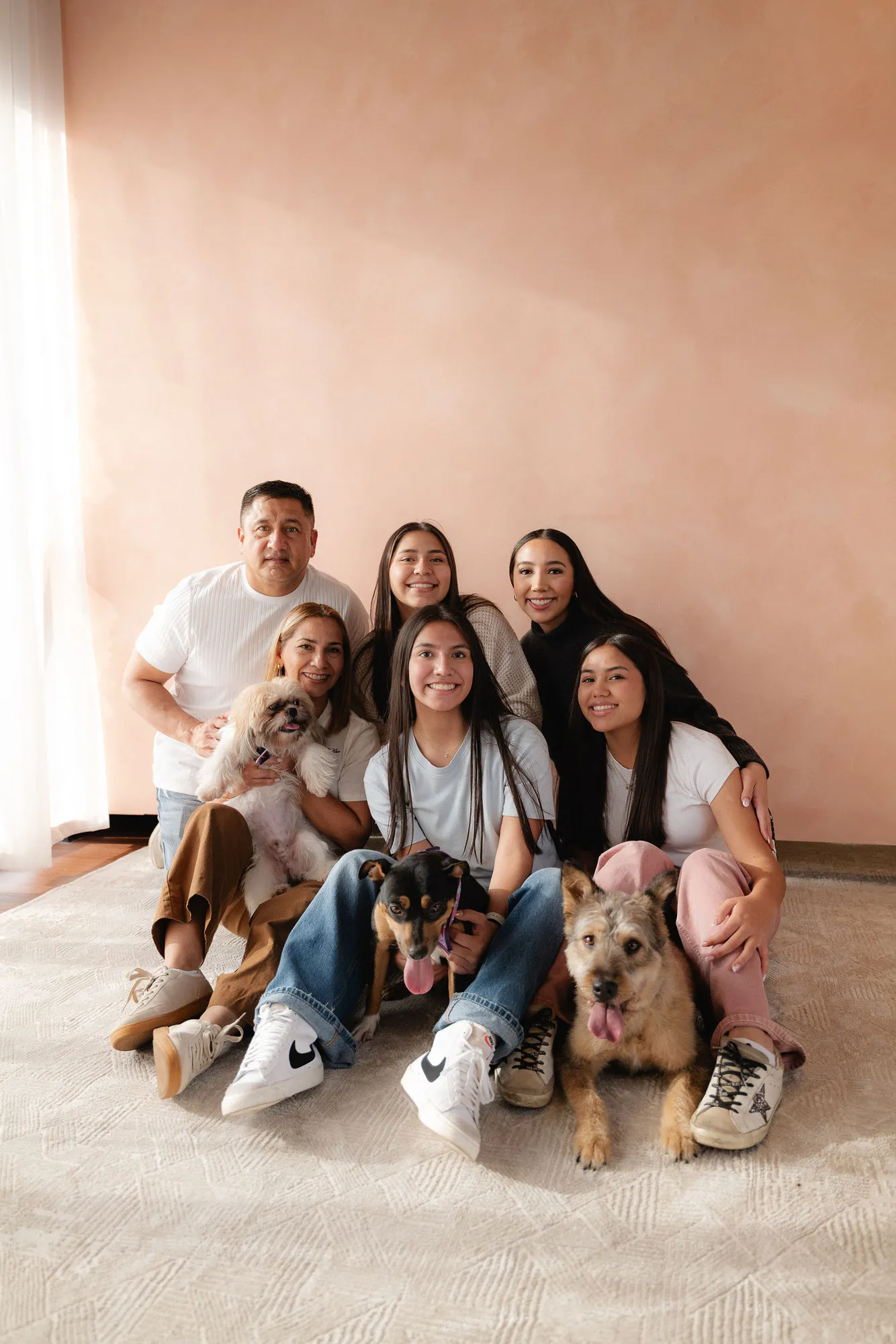 Una familia sonríe sentada en el piso junto a dos perros, frente a una pared color durazno.