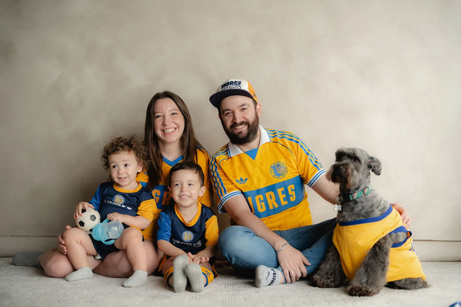 Una familia con jerseys amarillos de futbol y un perro posa sentada sobre un fondo claro.