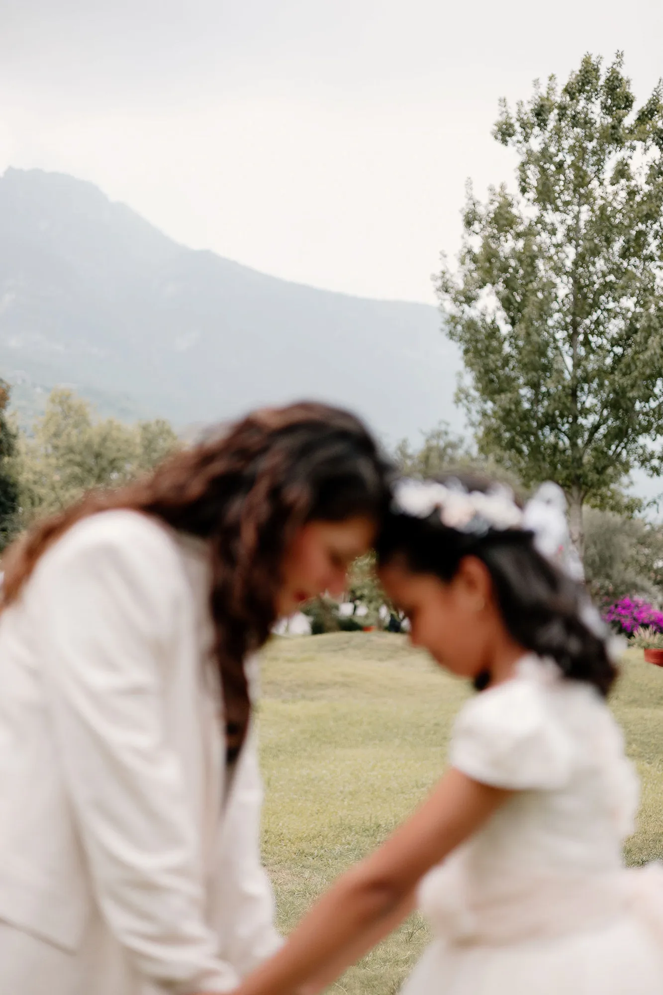 Una mamá y su hija se tocan la frente en un jardín; la niña lleva un vestido blanco y una diadema de flores.