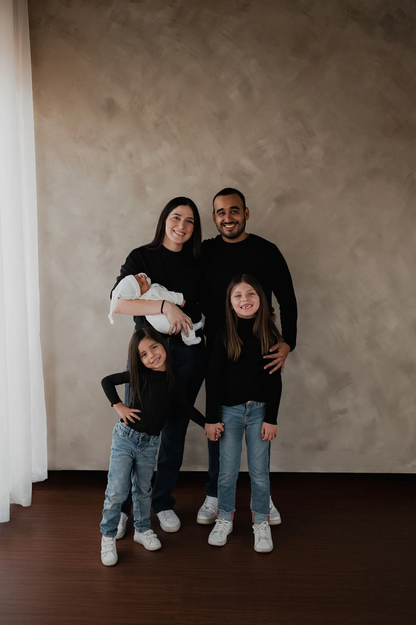 Una familia con un bebé posa de pie frente a una pared color arena.