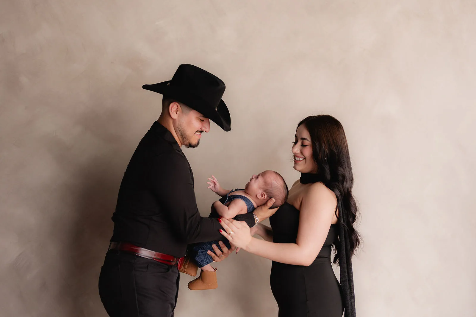 Un papá con sombrero vaquero y una mamá sostienen a su bebé frente a un fondo claro.