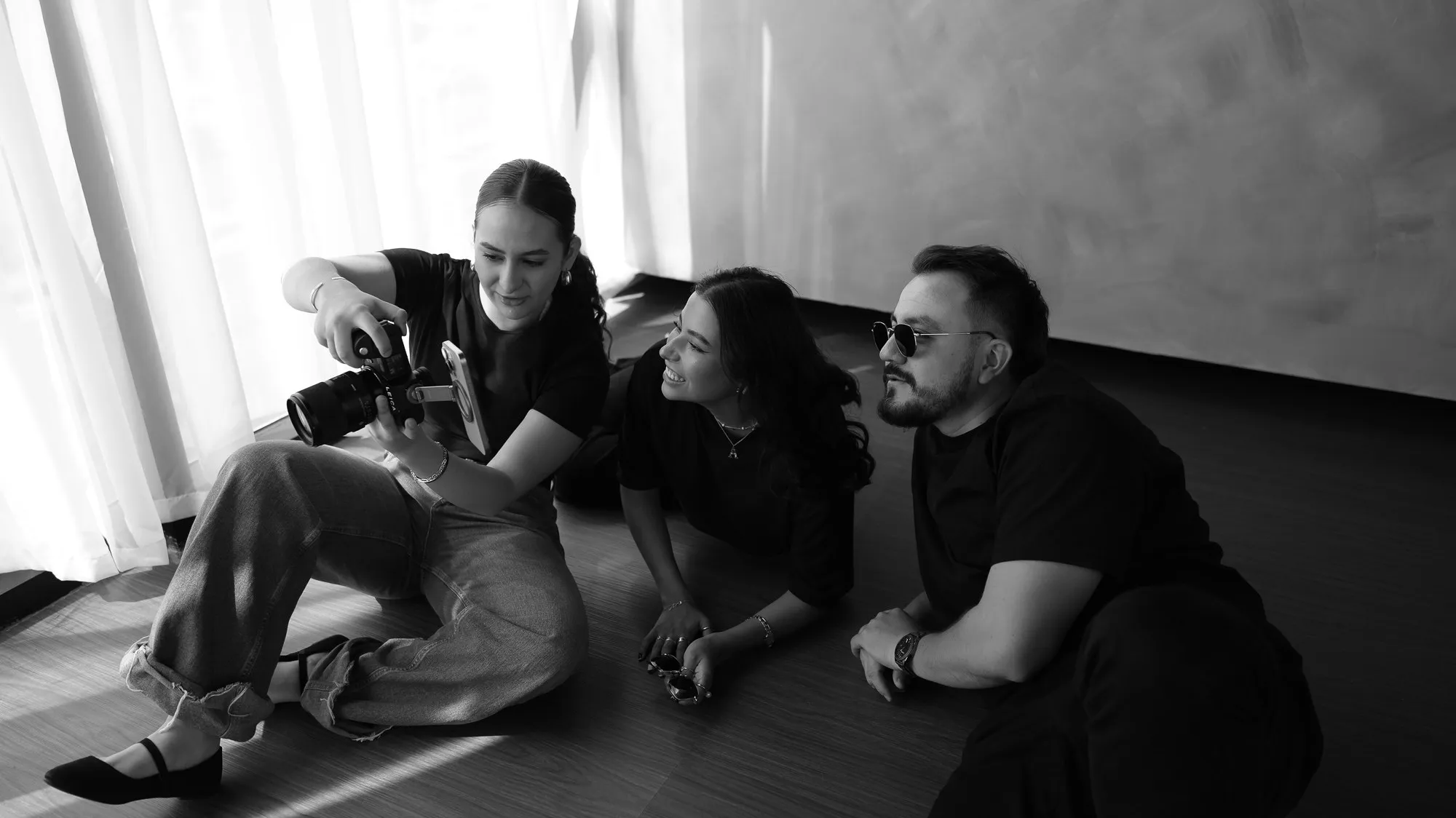 En blanco y negro, una fotógrafa sentada en el piso del estudio muestra una foto en su cámara a una mujer y a un hombre con lentes, que sonríen.