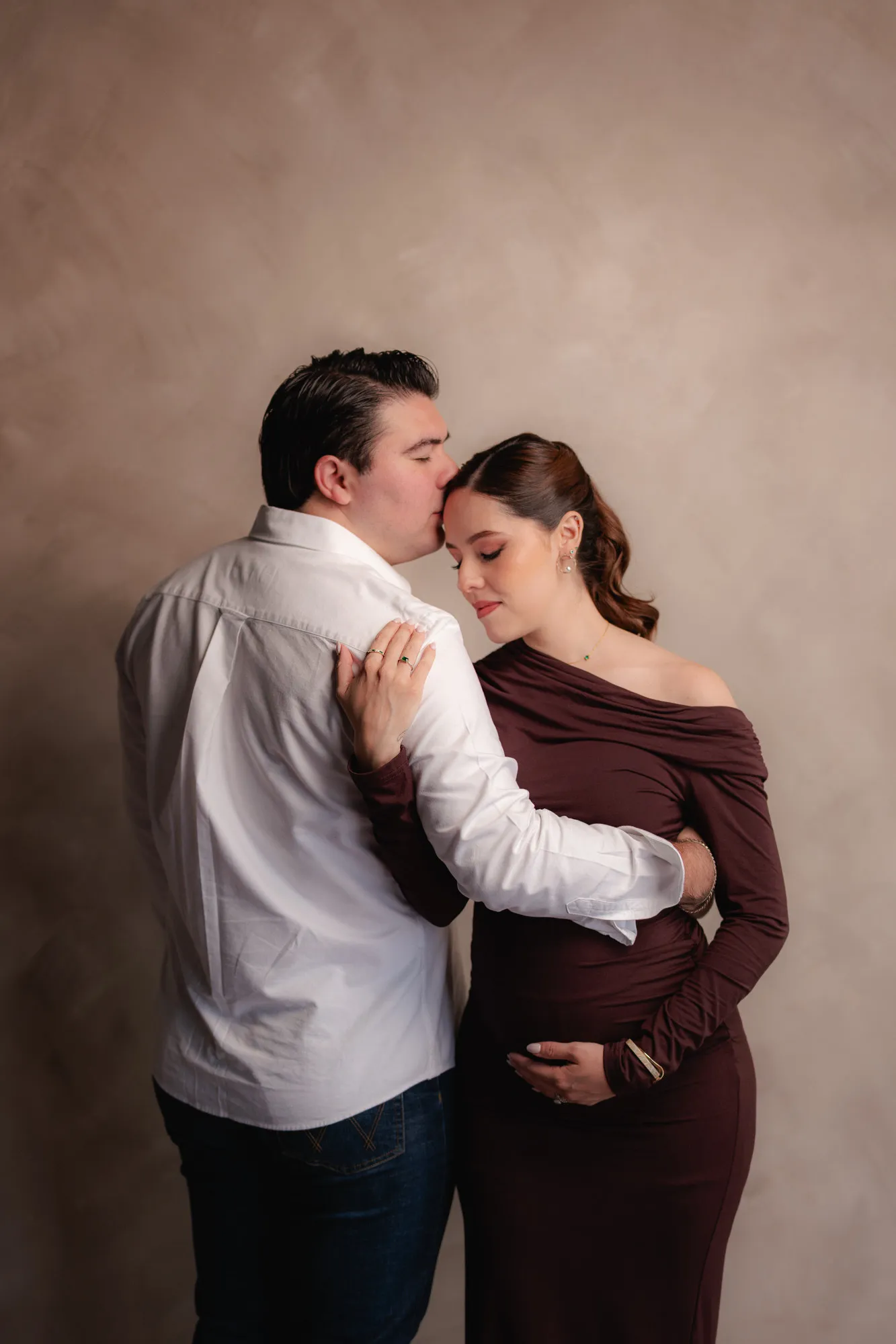 Una pareja se abraza; él besa la sien de ella, que lleva un vestido guinda y sostiene su vientre.