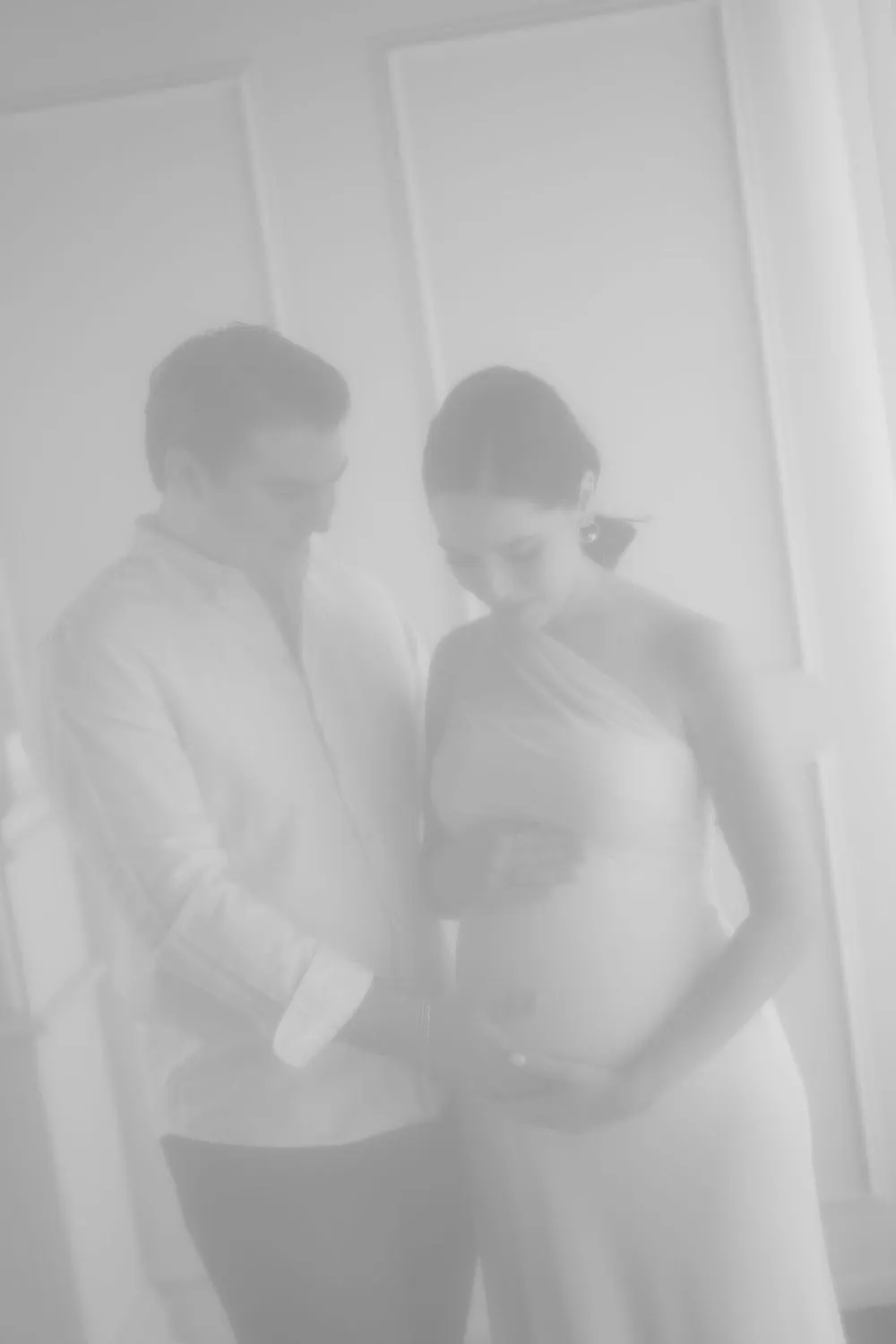 En blanco y negro, imagen suave y desvanecida de una pareja que abraza el vientre de ella.
