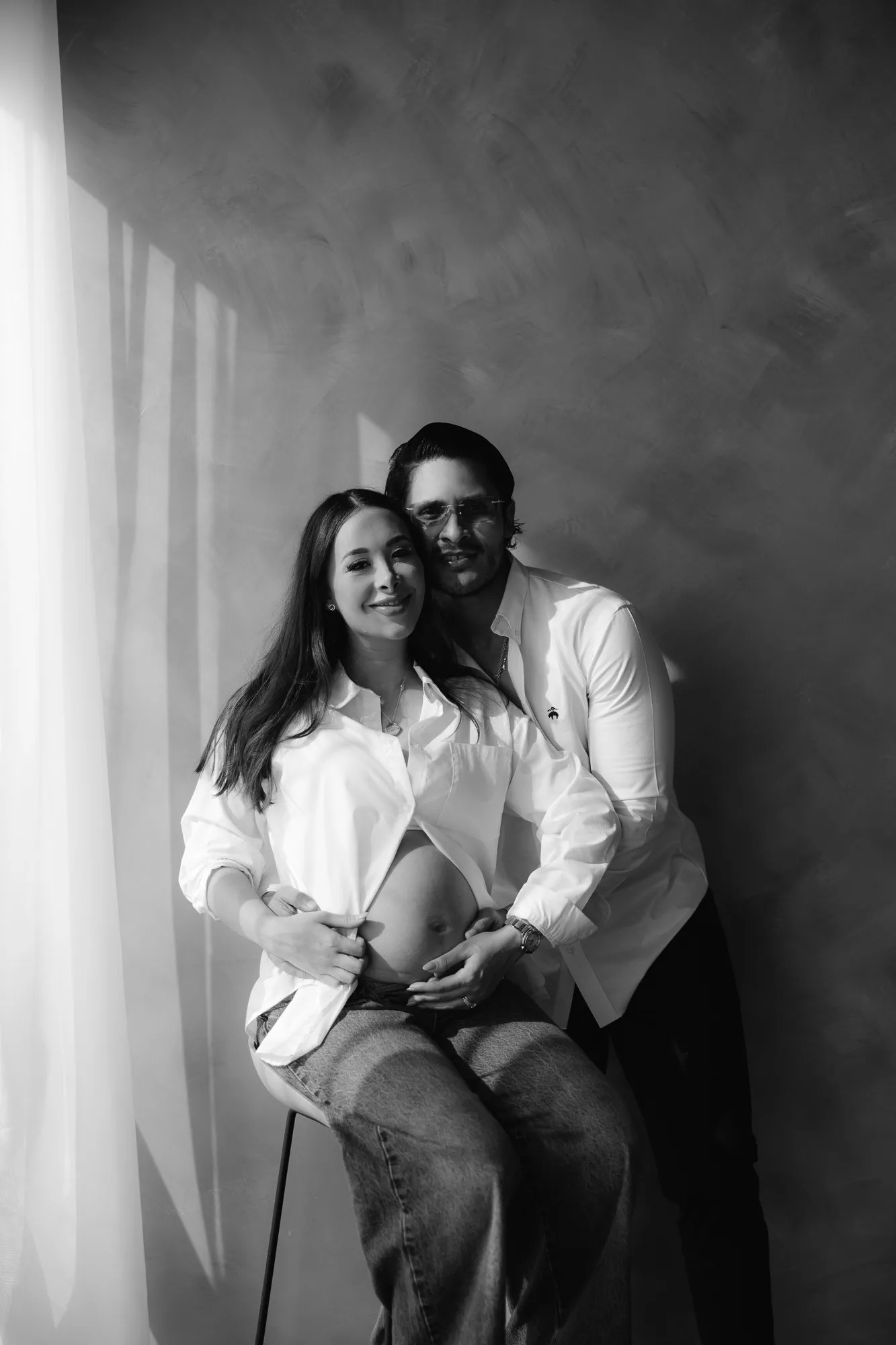 En blanco y negro, una pareja sentada se abraza; él apoya las manos en el vientre de ella.