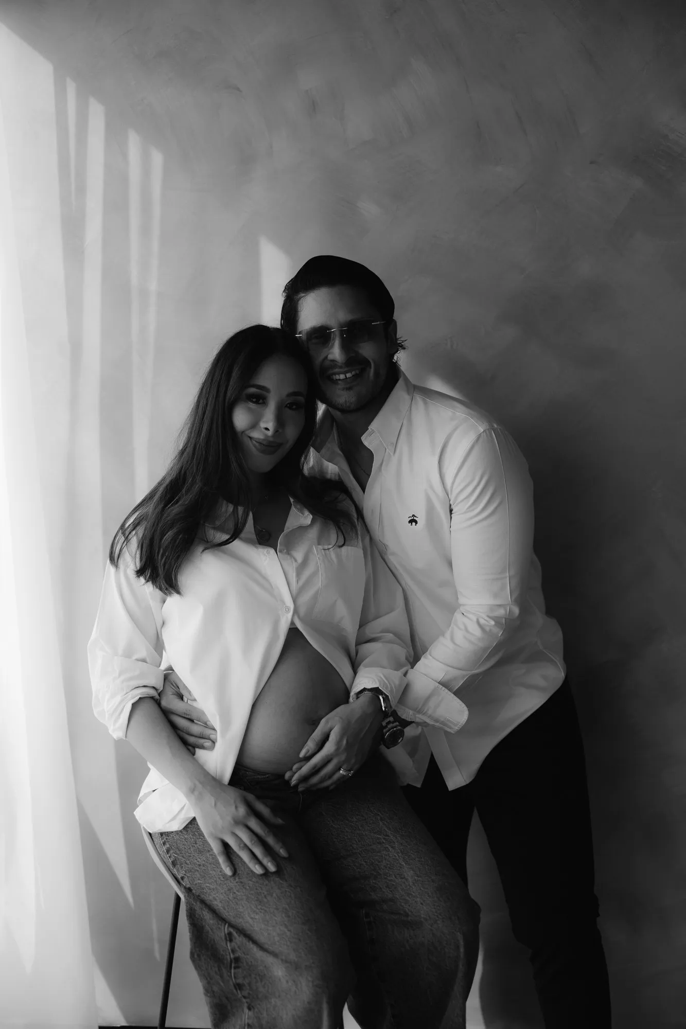 En blanco y negro, una pareja se abraza y sostiene el vientre de ella, junto a una cortina.