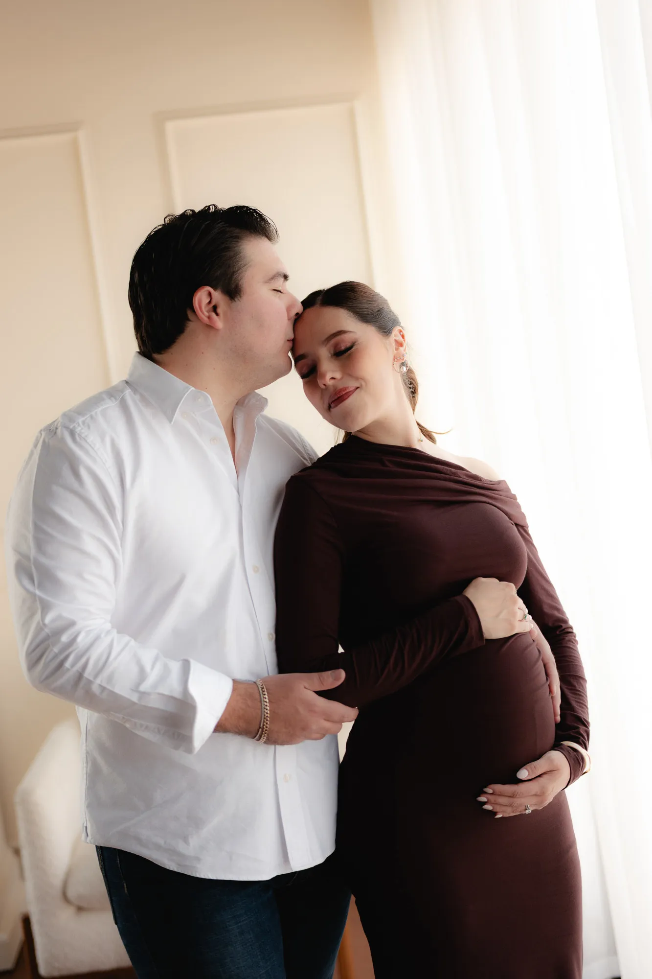 Una pareja se abraza; él besa la frente de ella, que lleva vestido guinda y sostiene su vientre.