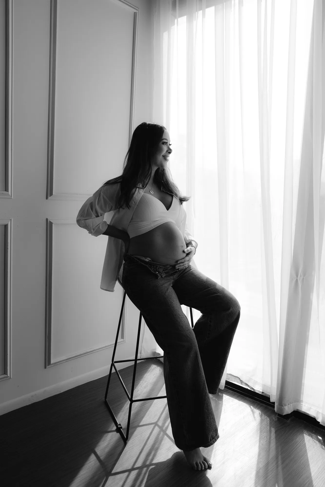 En blanco y negro, una mujer embarazada sentada en un banco alto junto a un ventanal.