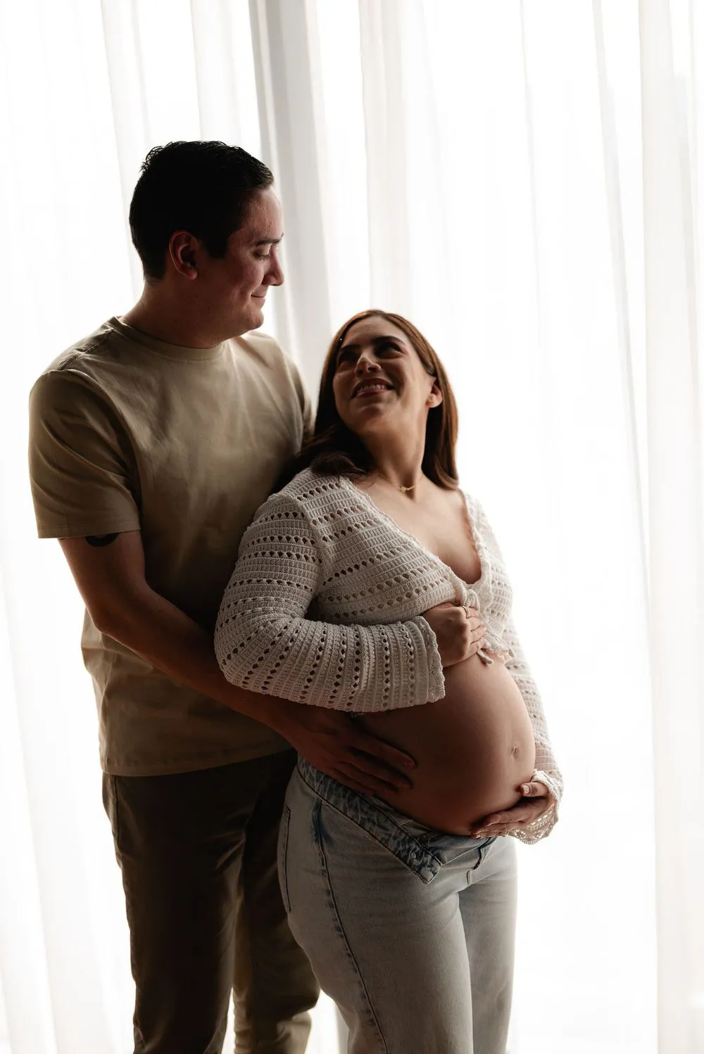 Una pareja se abraza de pie frente a cortinas blancas; ella apoya las manos en su vientre.