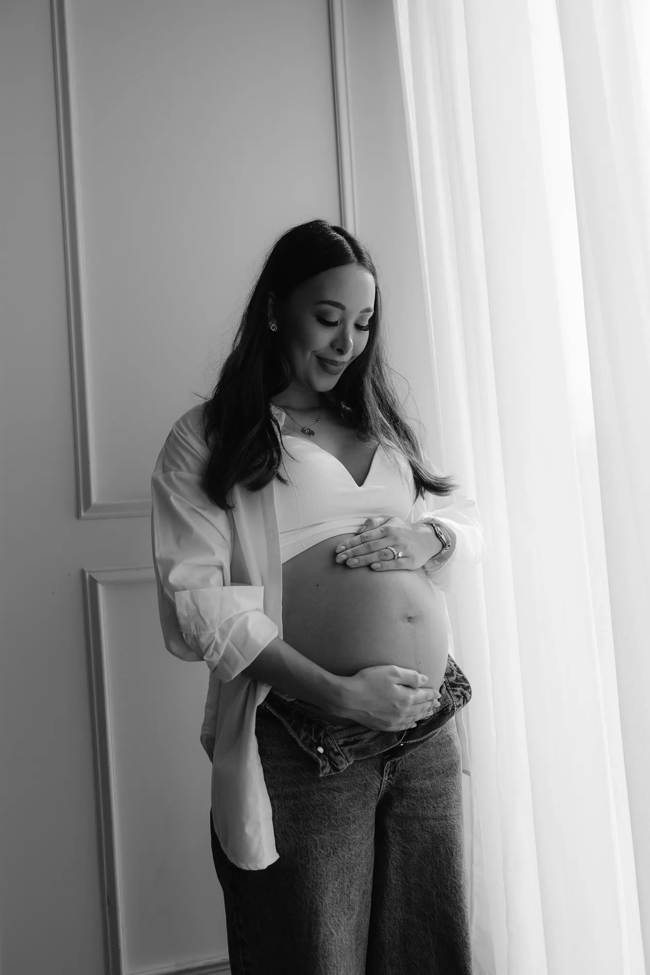 En blanco y negro, una mujer embarazada de pie junto a una cortina, con la camisa abierta.