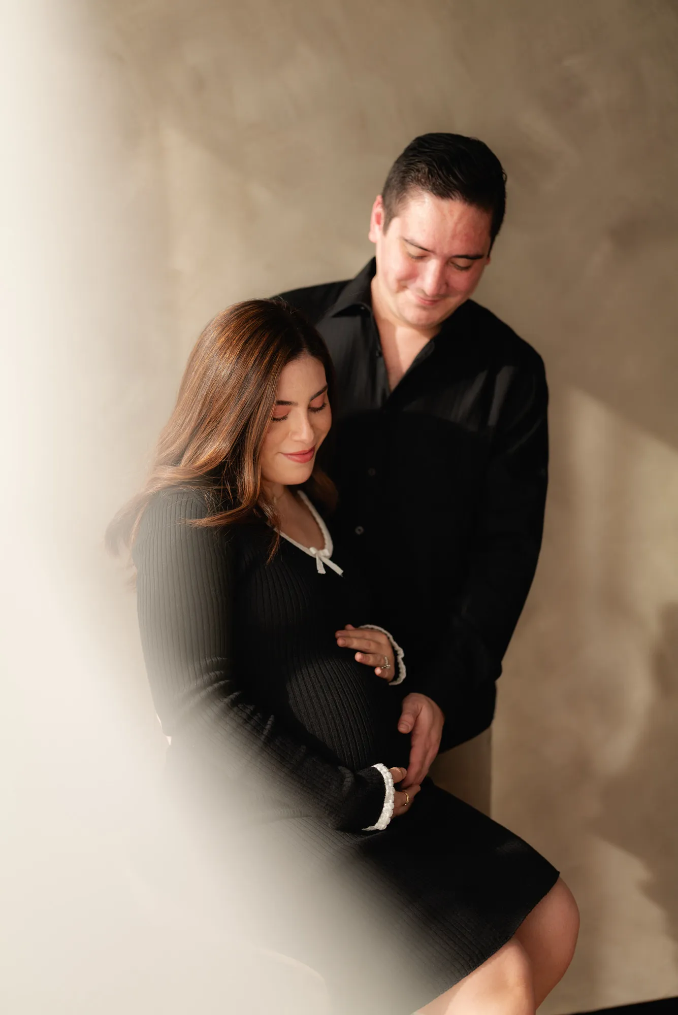 Una pareja sentada se abraza; ella viste de negro y él, camisa oscura, frente a una pared color arena.
