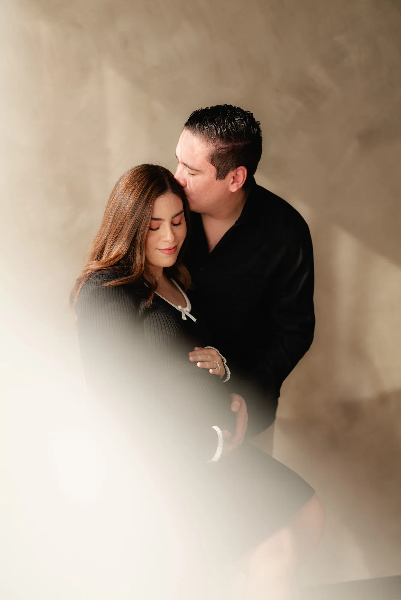 Una pareja se abraza; ella viste de negro y una luz difusa cae a un lado, frente a una pared color arena.