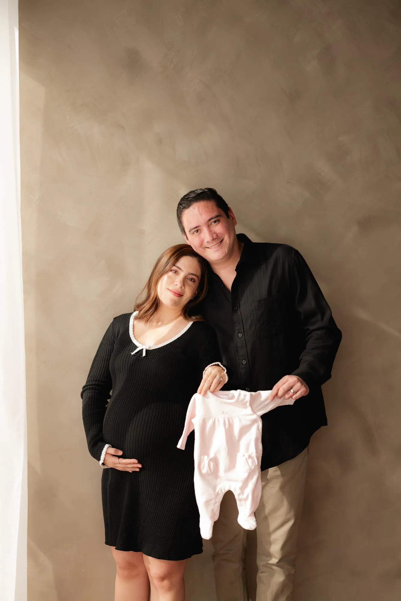 Una pareja posa abrazada; ella sostiene un conjunto rosa de bebé.