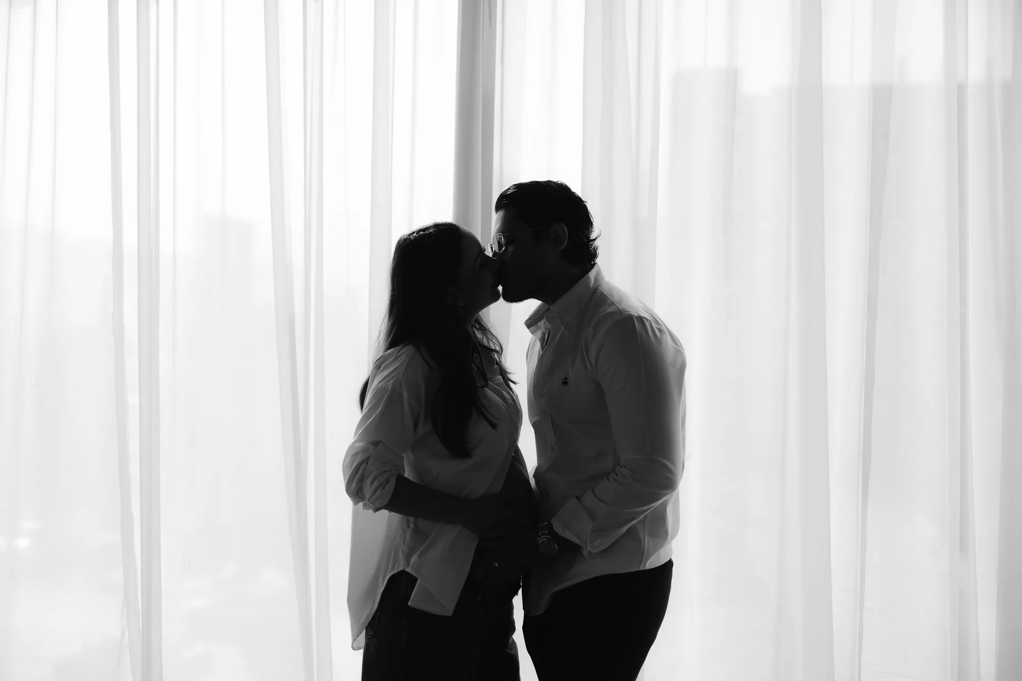 En blanco y negro, una pareja se besa a contraluz frente a ventanales con cortinas.
