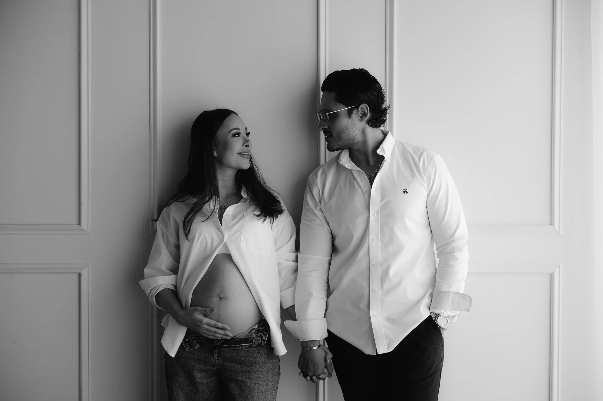 En blanco y negro, una pareja de pie; él con camisa blanca y ella con la camisa abierta que muestra su vientre.