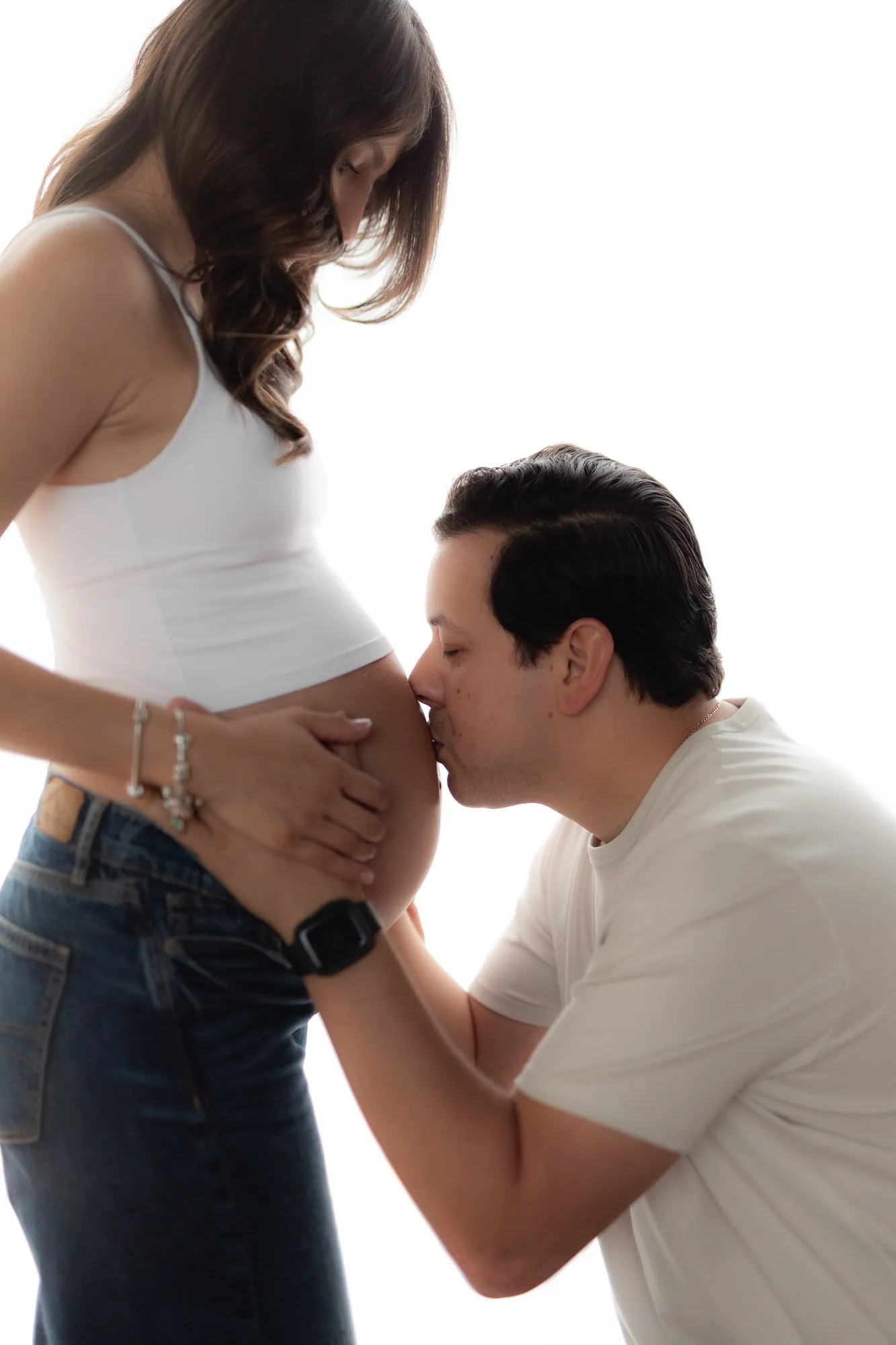 Él besa el vientre de su pareja, que lleva top blanco, sobre fondo blanco.
