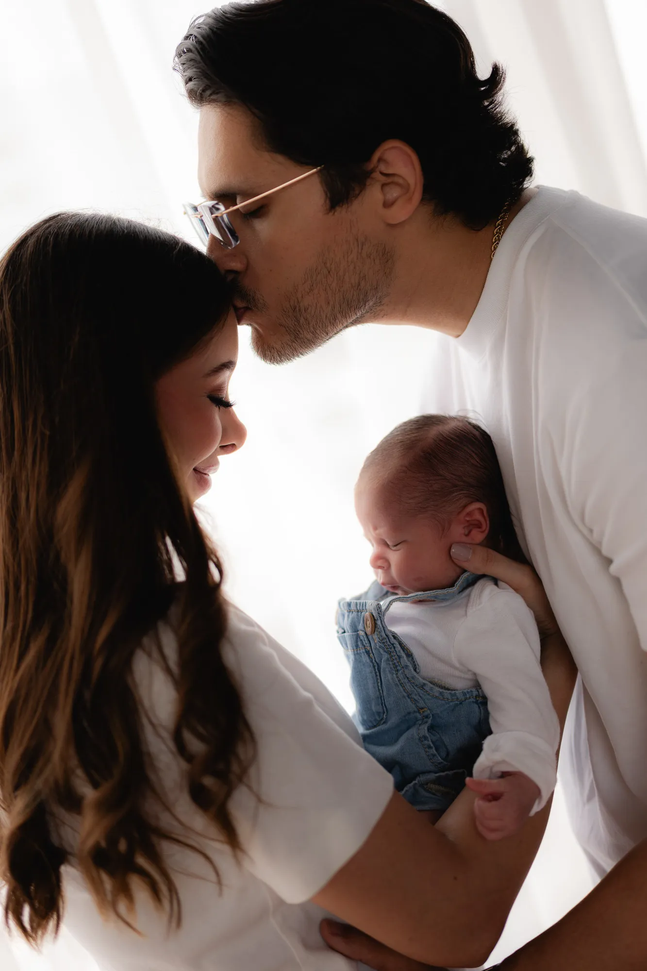 Un papá besa la frente de la mamá mientras ella sostiene a su bebé, a contraluz.