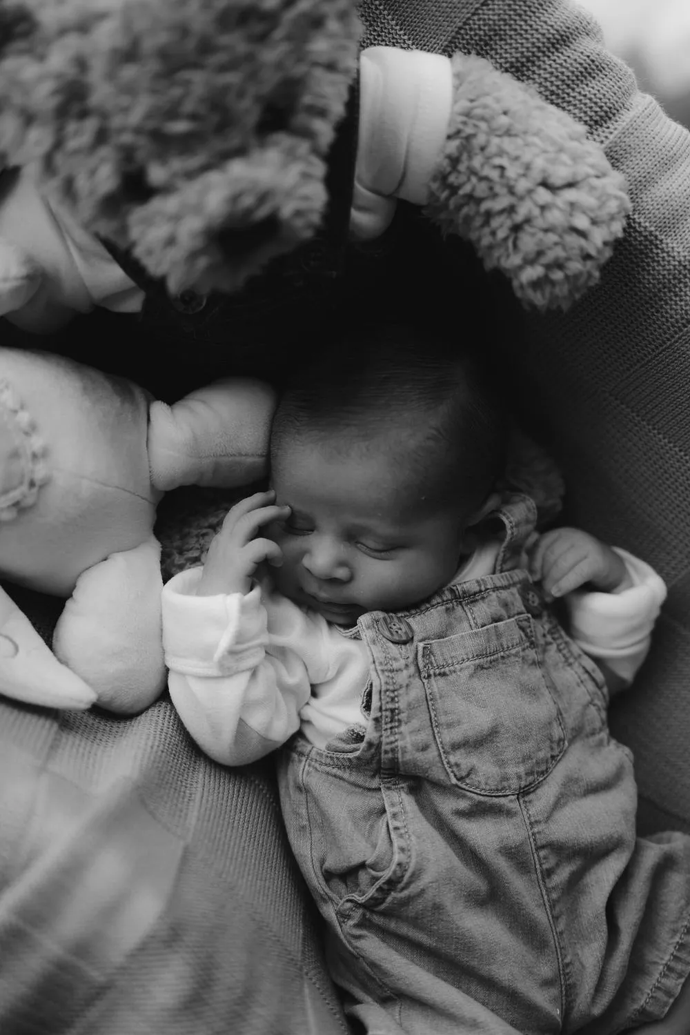 En blanco y negro, un bebé dormido sobre una cobija junto a un peluche.