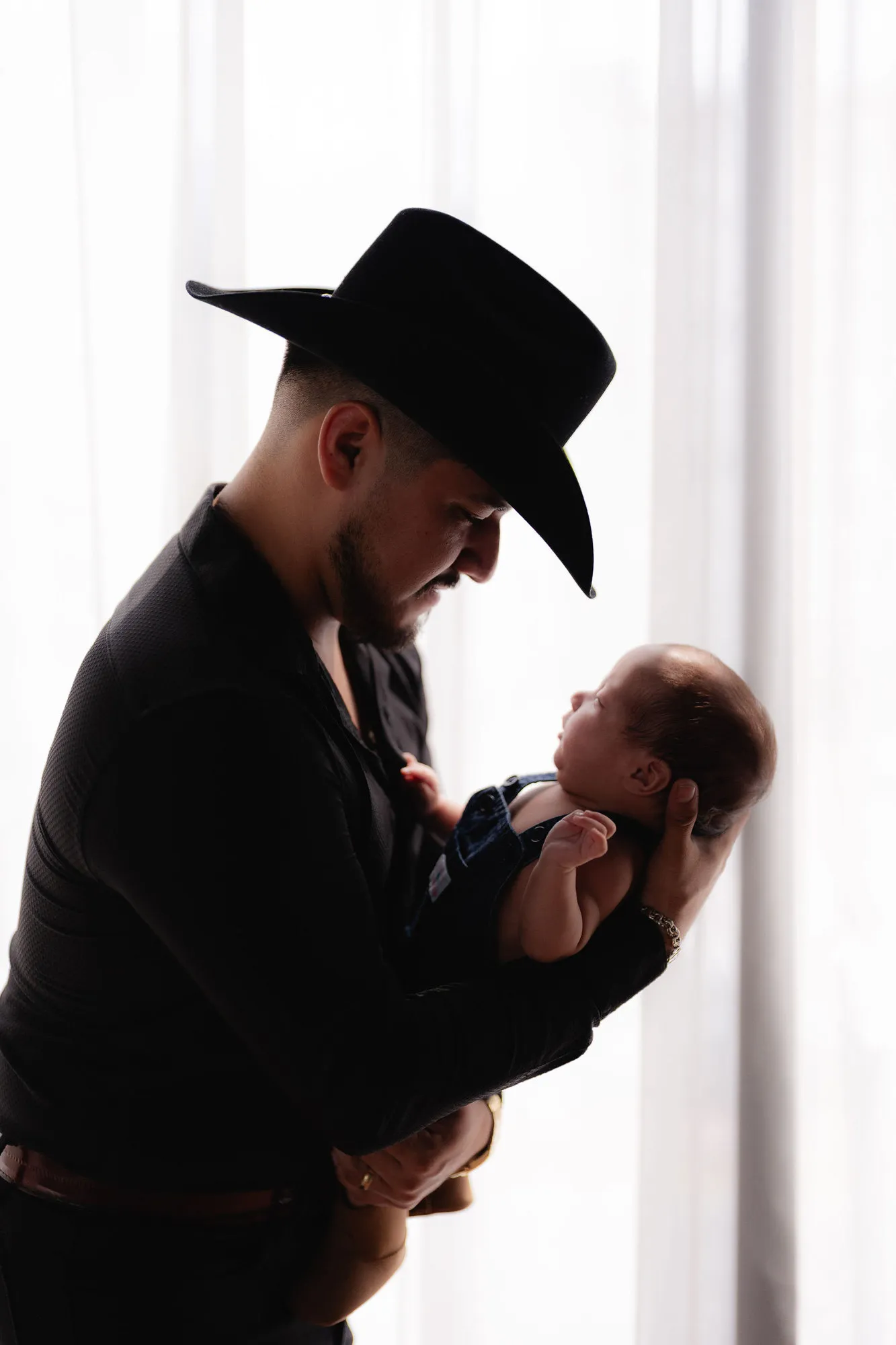 Un papá con sombrero vaquero sostiene a su bebé frente a una cortina blanca.