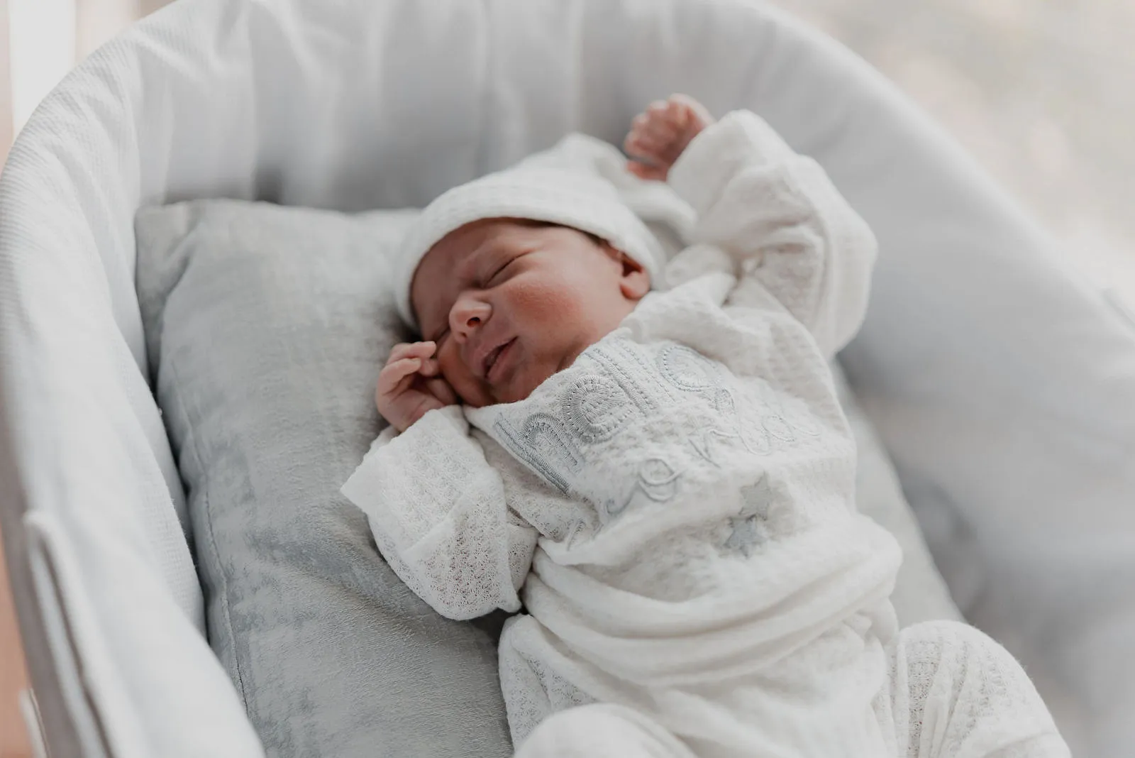 Un recién nacido dormido en un moisés gris, con gorro y pijama blanca.