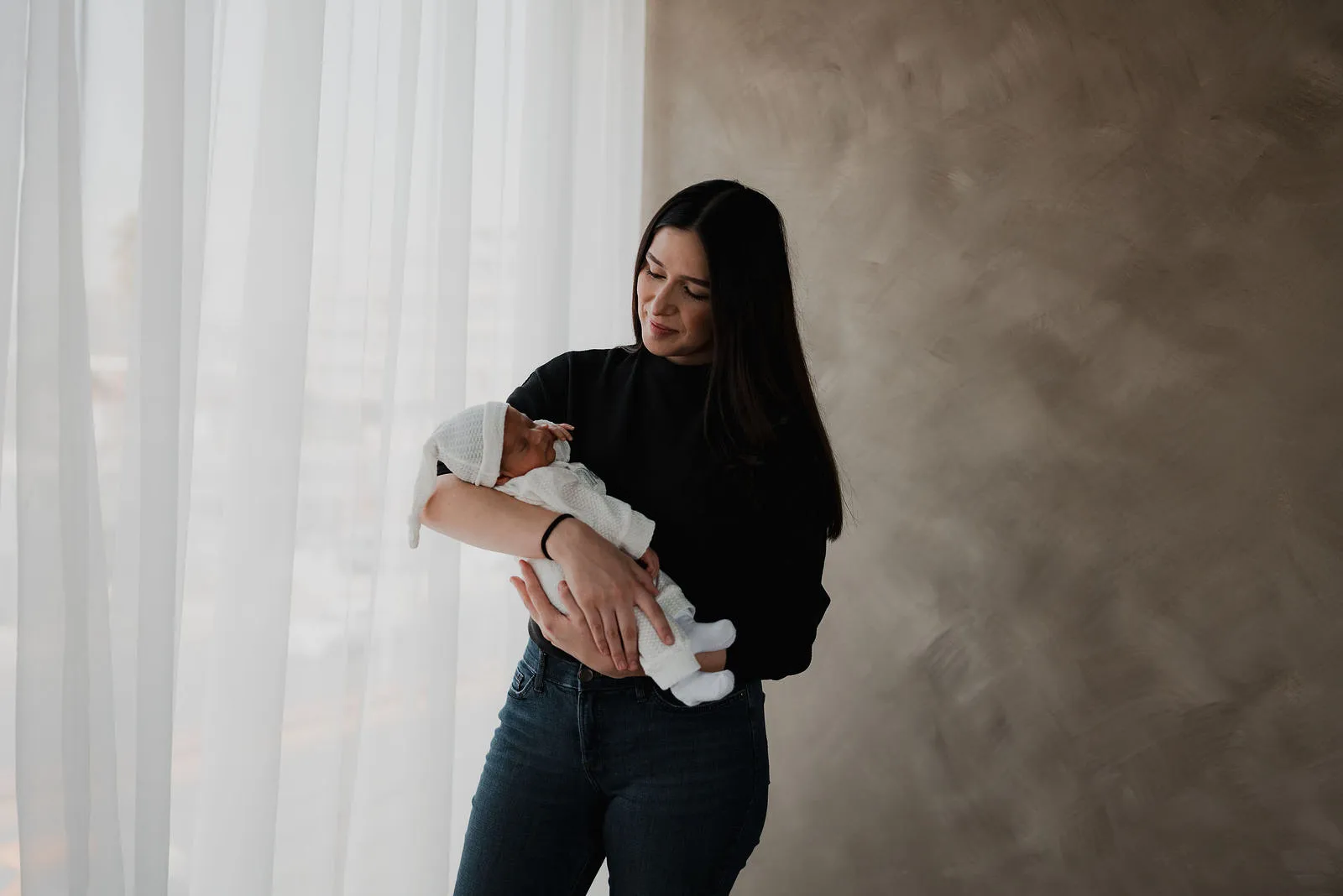 Una mamá de pie junto a un ventanal sostiene a su bebé; se ve de lejos.