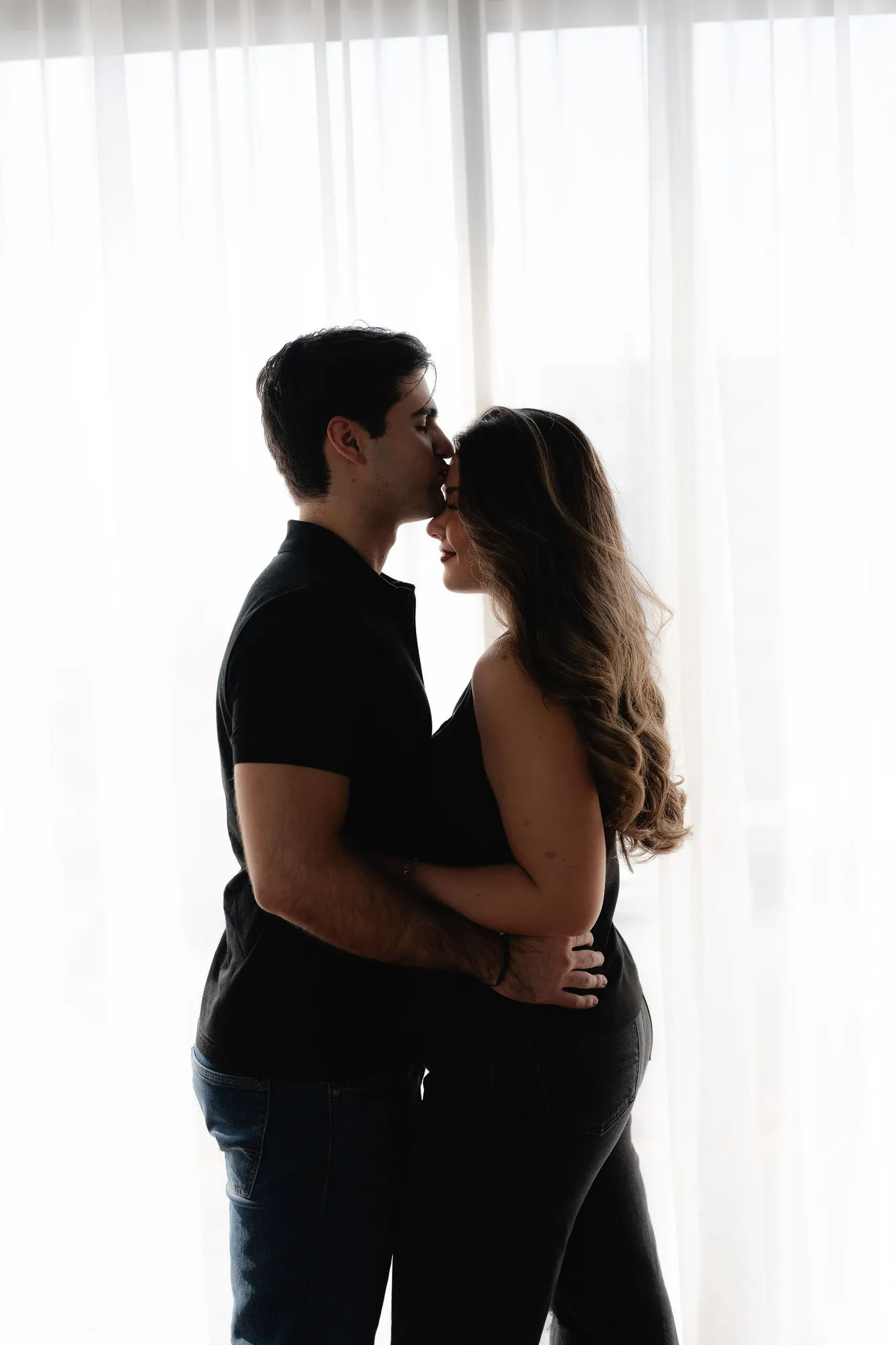 Una pareja se abraza frente a una cortina blanca; él le besa la frente.