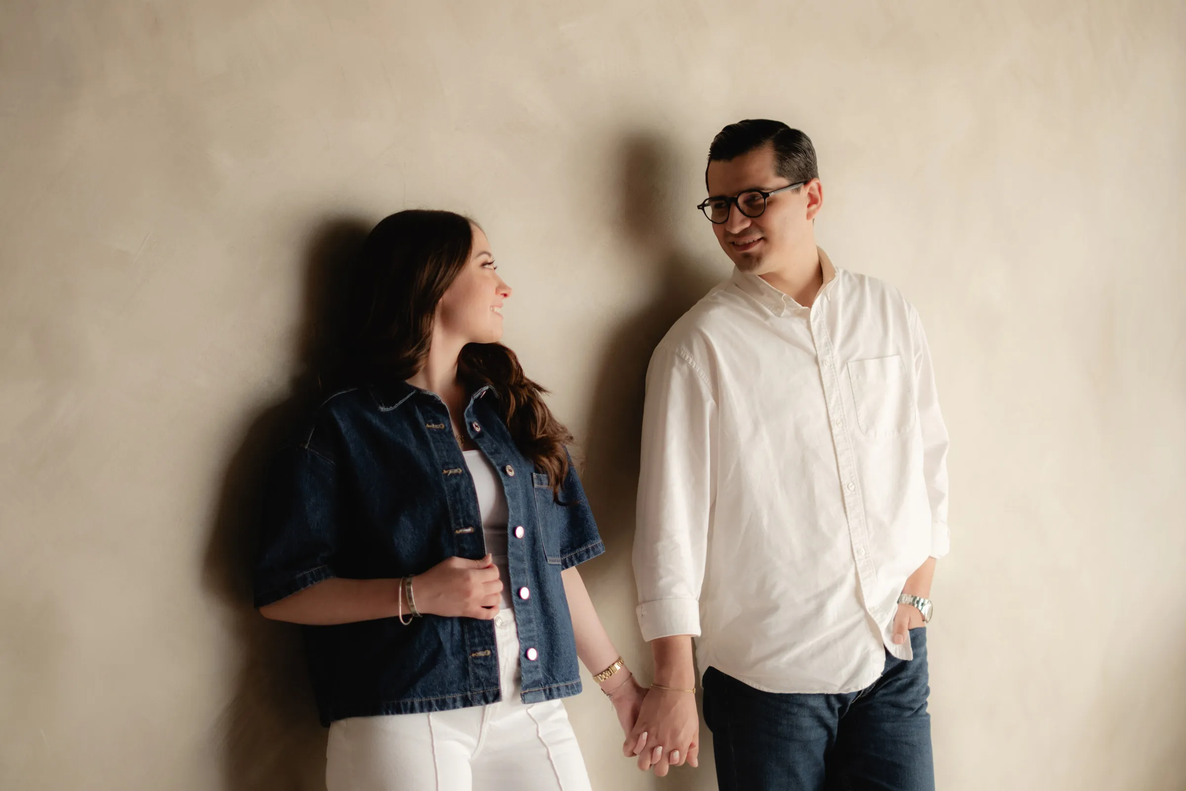 Una pareja se toma de la mano, recargada en una pared color arena: ella con chamarra de mezclilla y él con camisa blanca, se miran sonriendo.