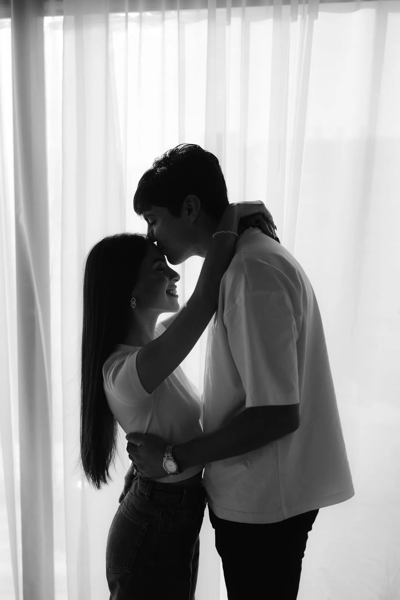 En blanco y negro, una pareja se besa entre cortinas blancas, a contraluz.
