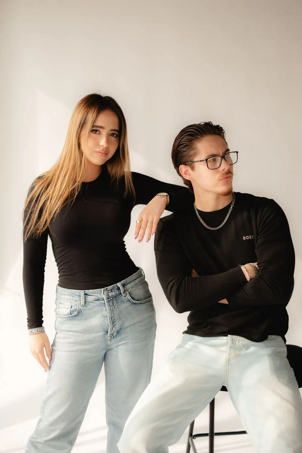 Una pareja posa con ropa negra y jeans claros; ella se apoya en él, que cruza los brazos.