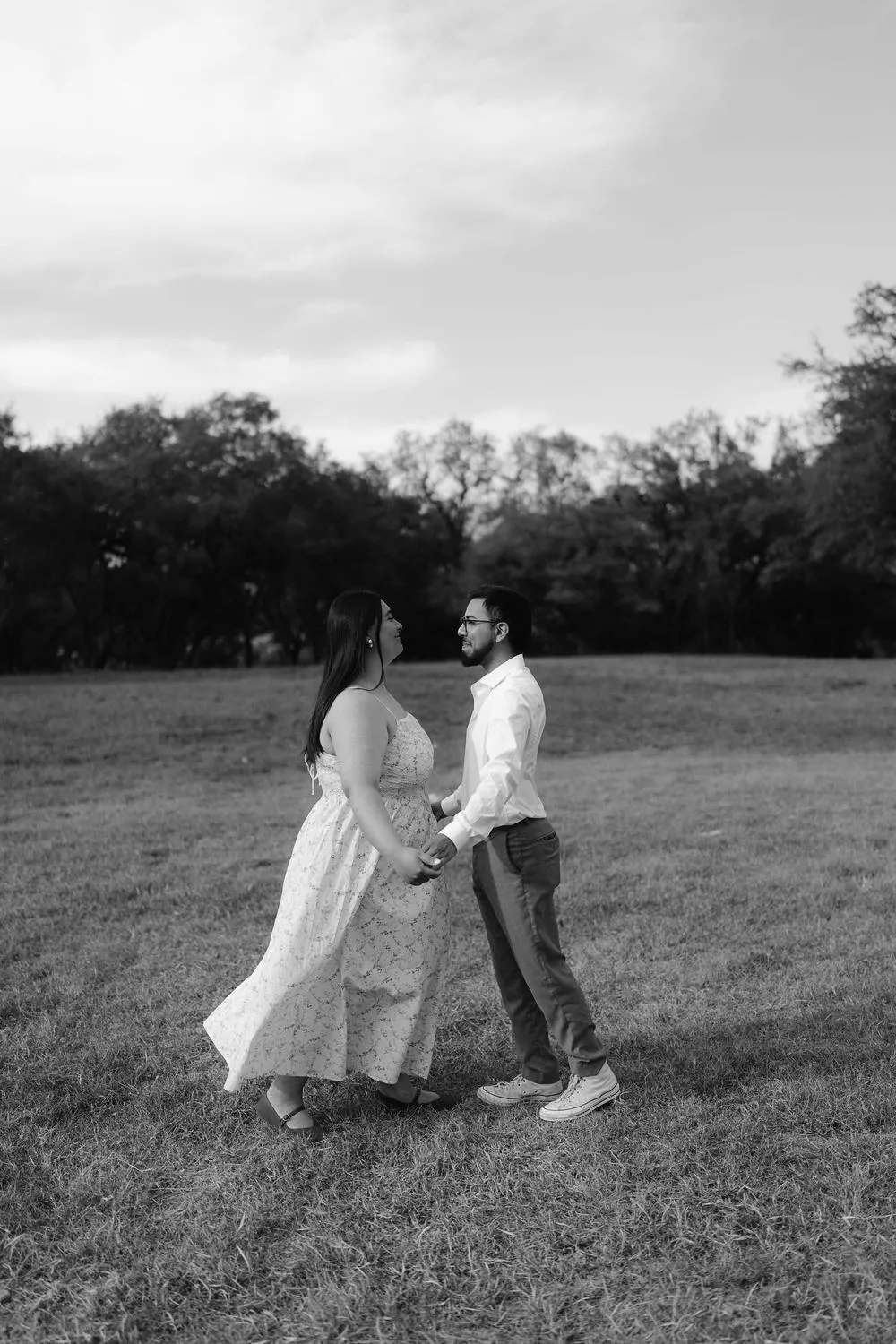 En blanco y negro, una pareja se abraza en un campo abierto, con árboles al fondo.