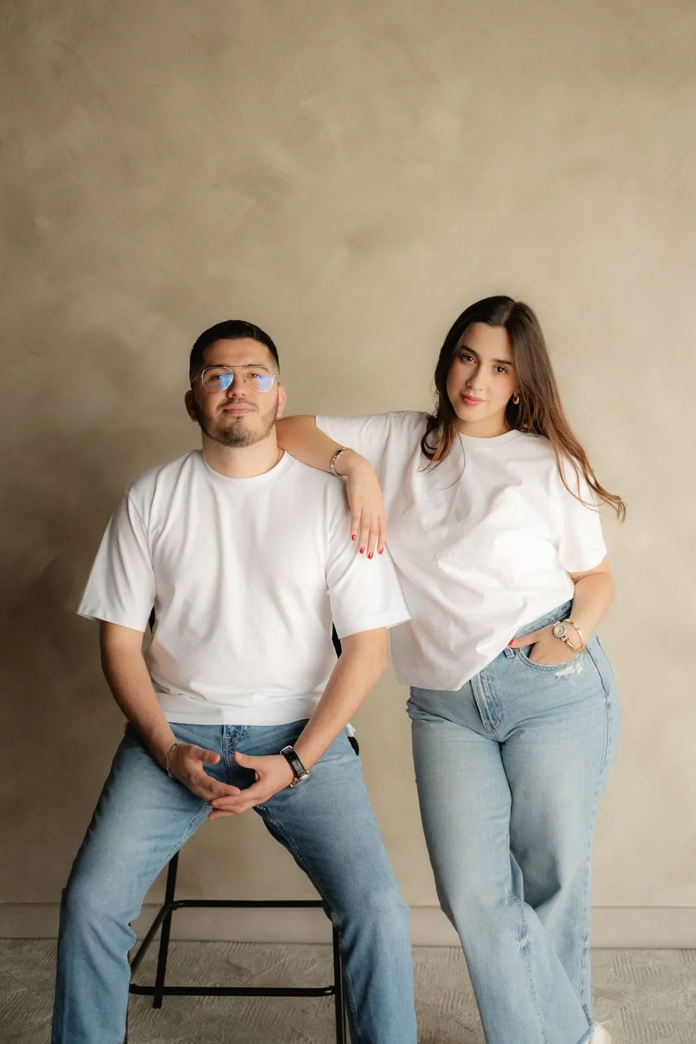 Una pareja sentada en un banco, con playera blanca y jeans, frente a una pared color arena.