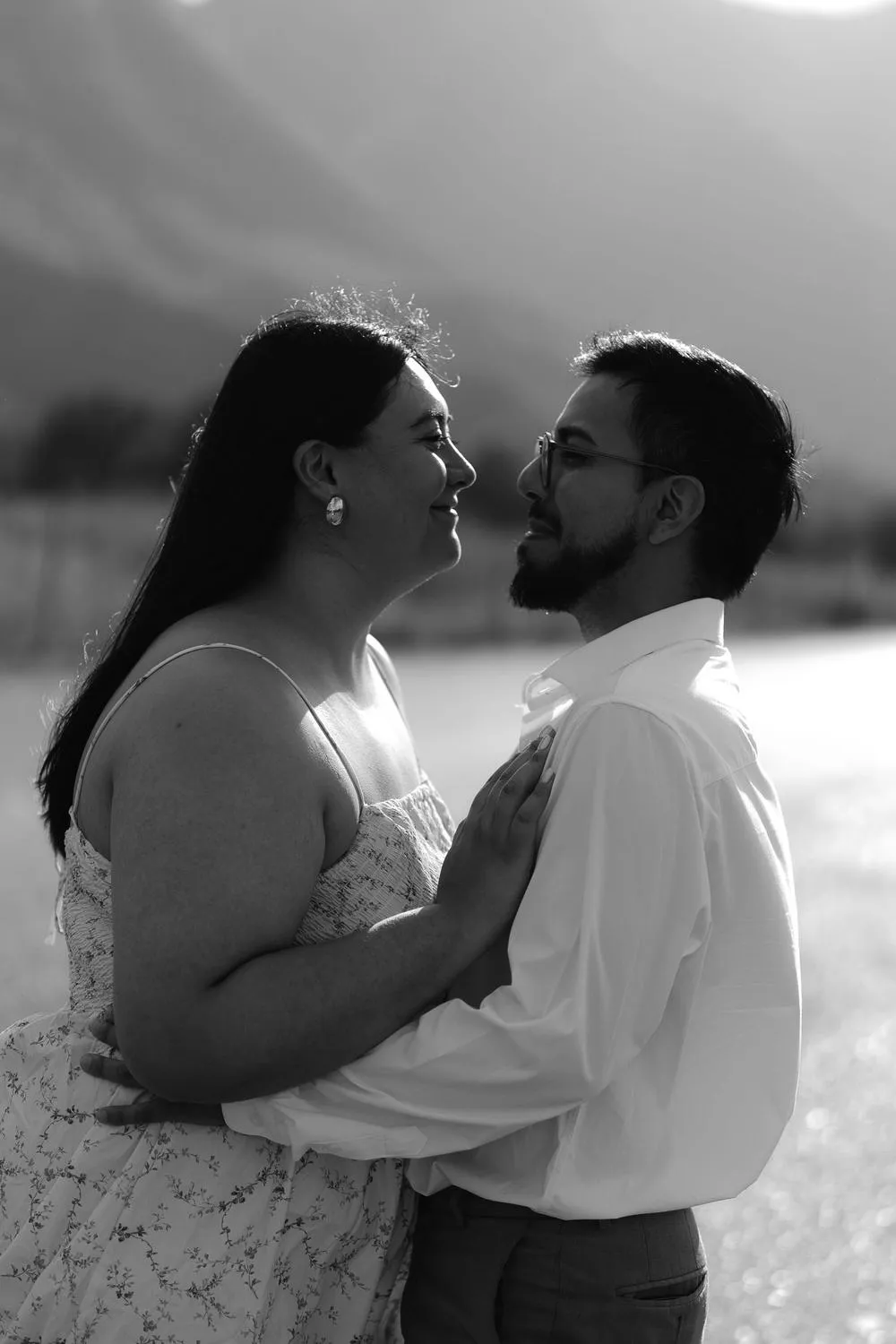En blanco y negro, una pareja se abraza y se mira de cerca, con montañas al fondo.