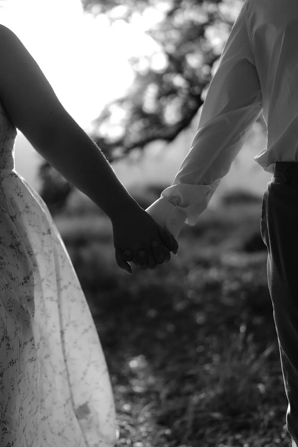 En blanco y negro, una pareja se toma de la mano en el campo; solo se ven sus manos y parte de su ropa.