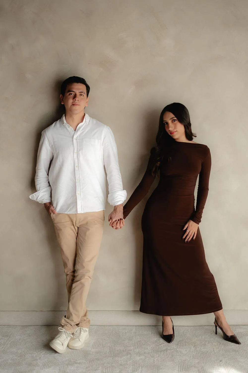 Una pareja de pie se toma de la mano, recargada en una pared color arena; él de camisa blanca y ella de vestido largo café.