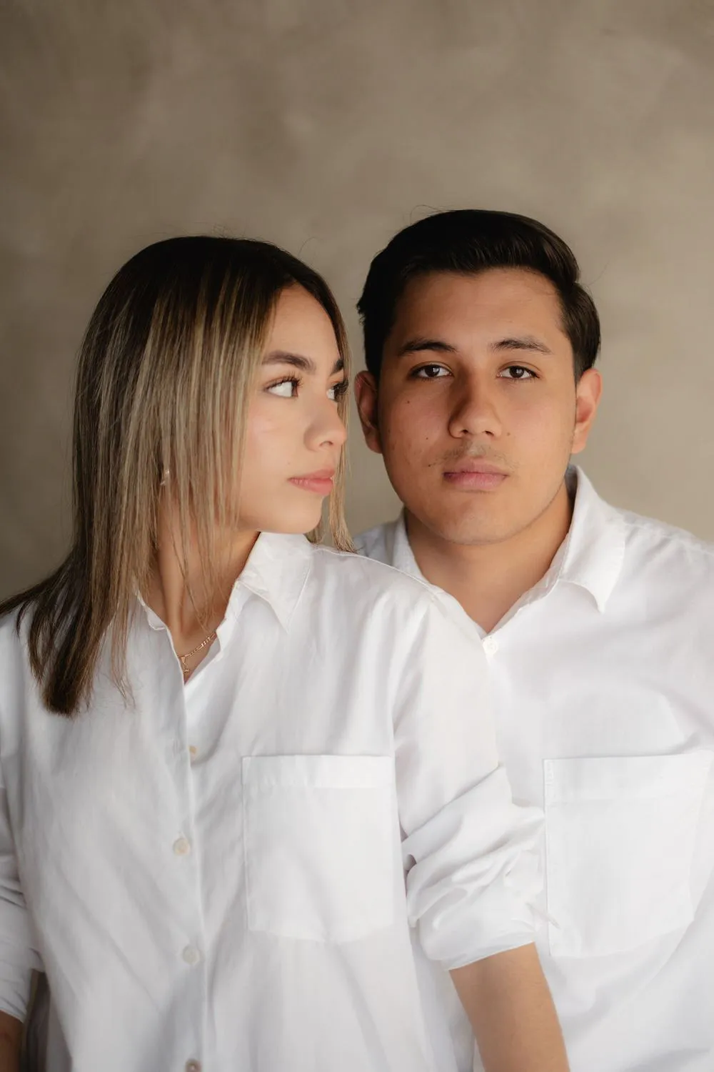 Retrato de una pareja con camisas blancas que mira a la cámara, frente a una pared color arena.