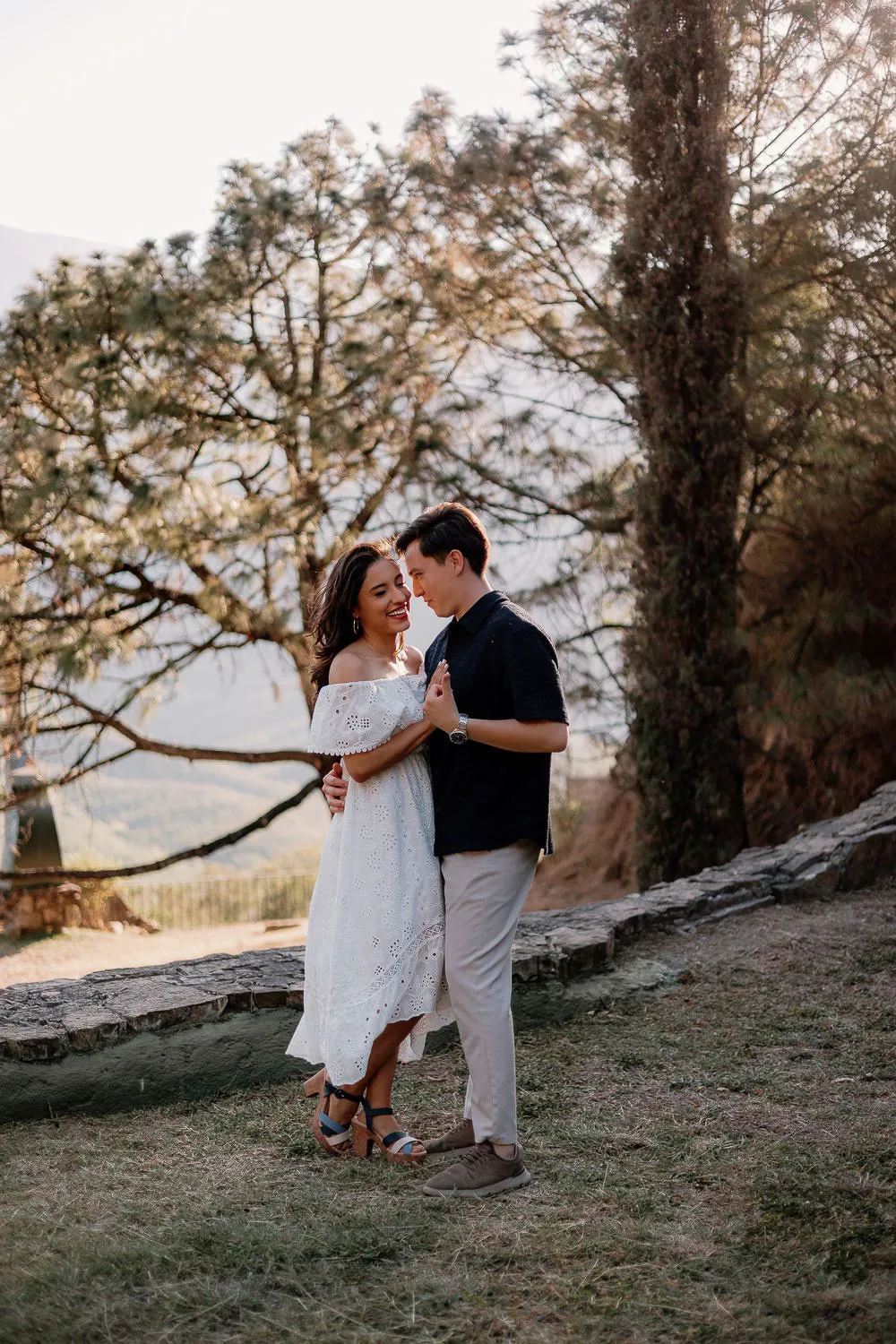 Una pareja se abraza en un mirador entre pinos, con vestido blanco y camisa oscura.
