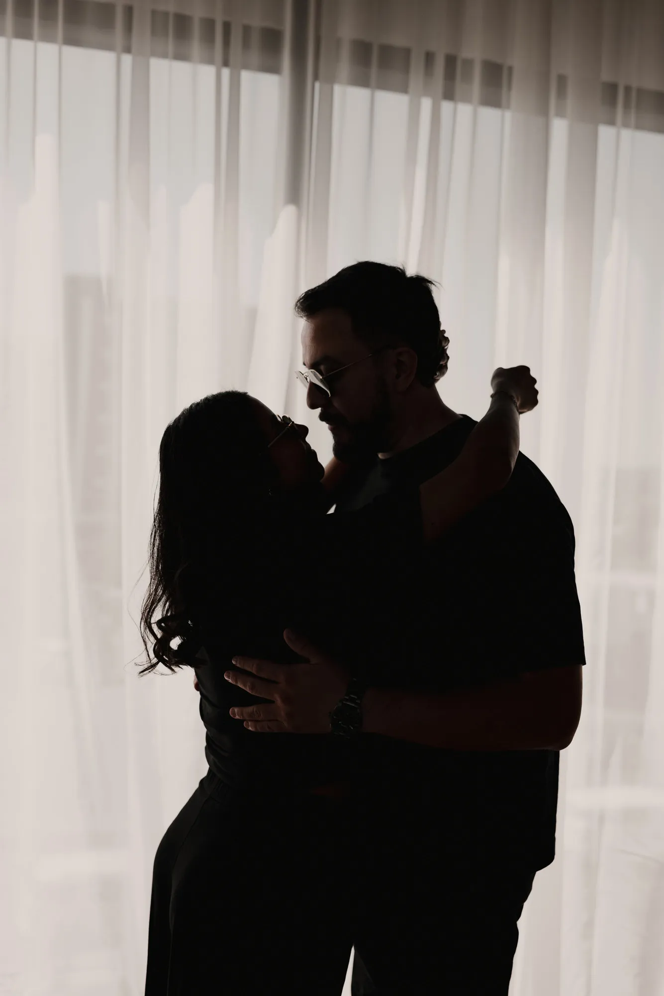 En blanco y negro, una pareja se abraza a contraluz frente a cortinas de gasa.