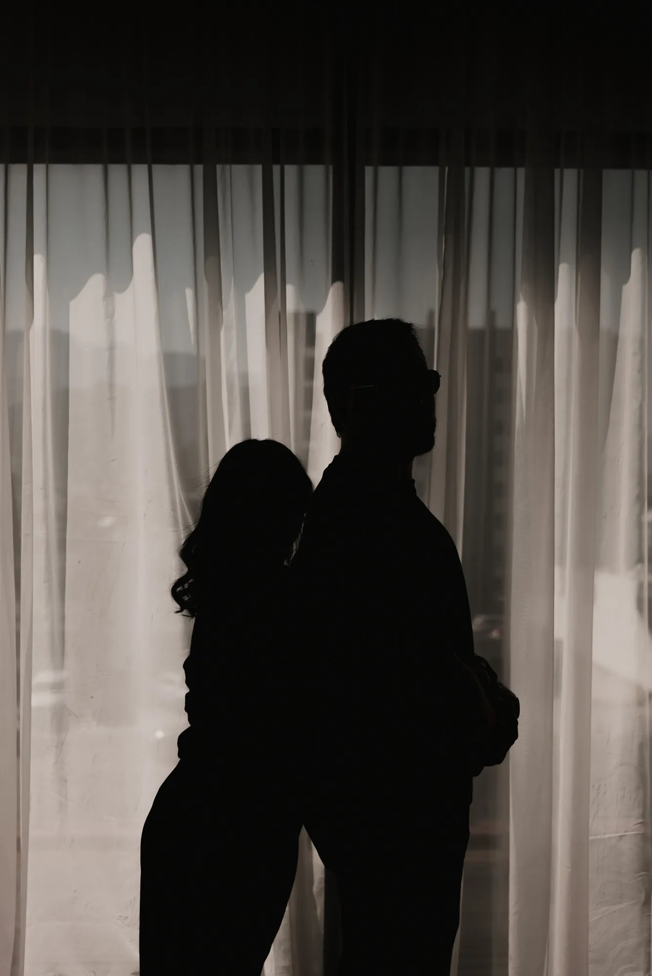 En blanco y negro, las siluetas de una pareja de pie frente a cortinas transparentes.