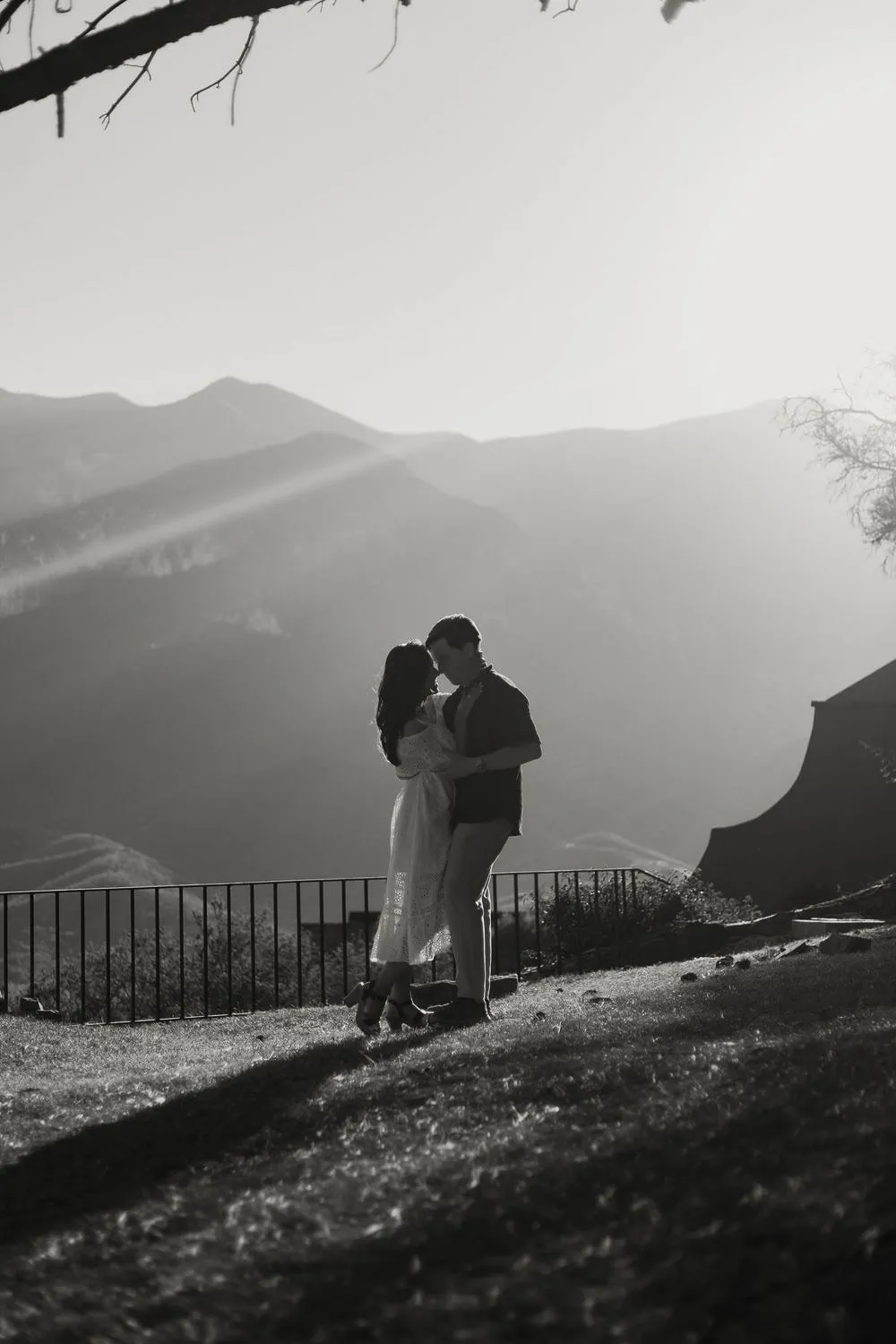 En blanco y negro, una pareja se abraza a lo lejos en un mirador, con montañas y luz de atardecer.