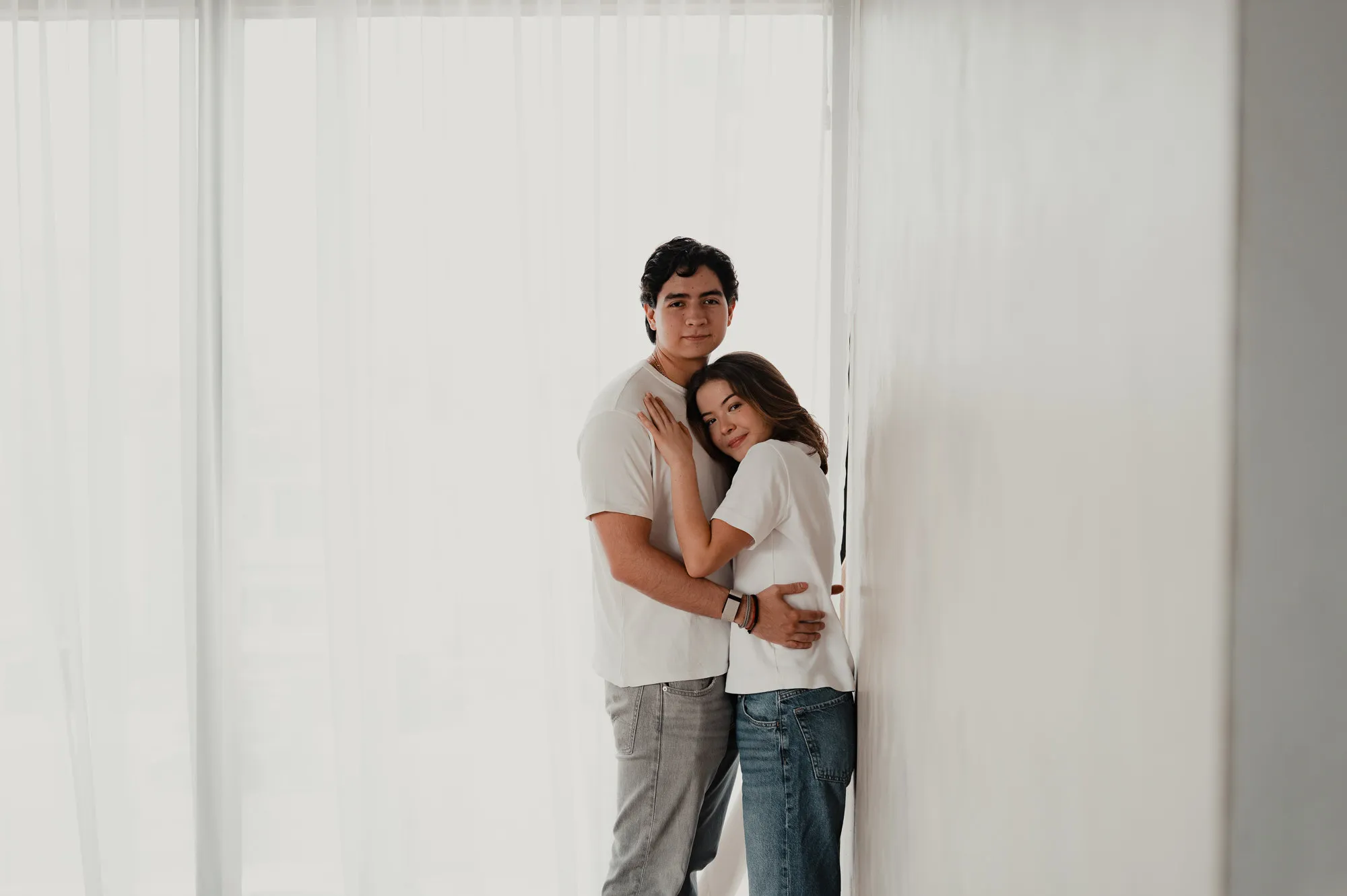 Una pareja se abraza de pie frente a una cortina blanca; se ven pequeños en la imagen.