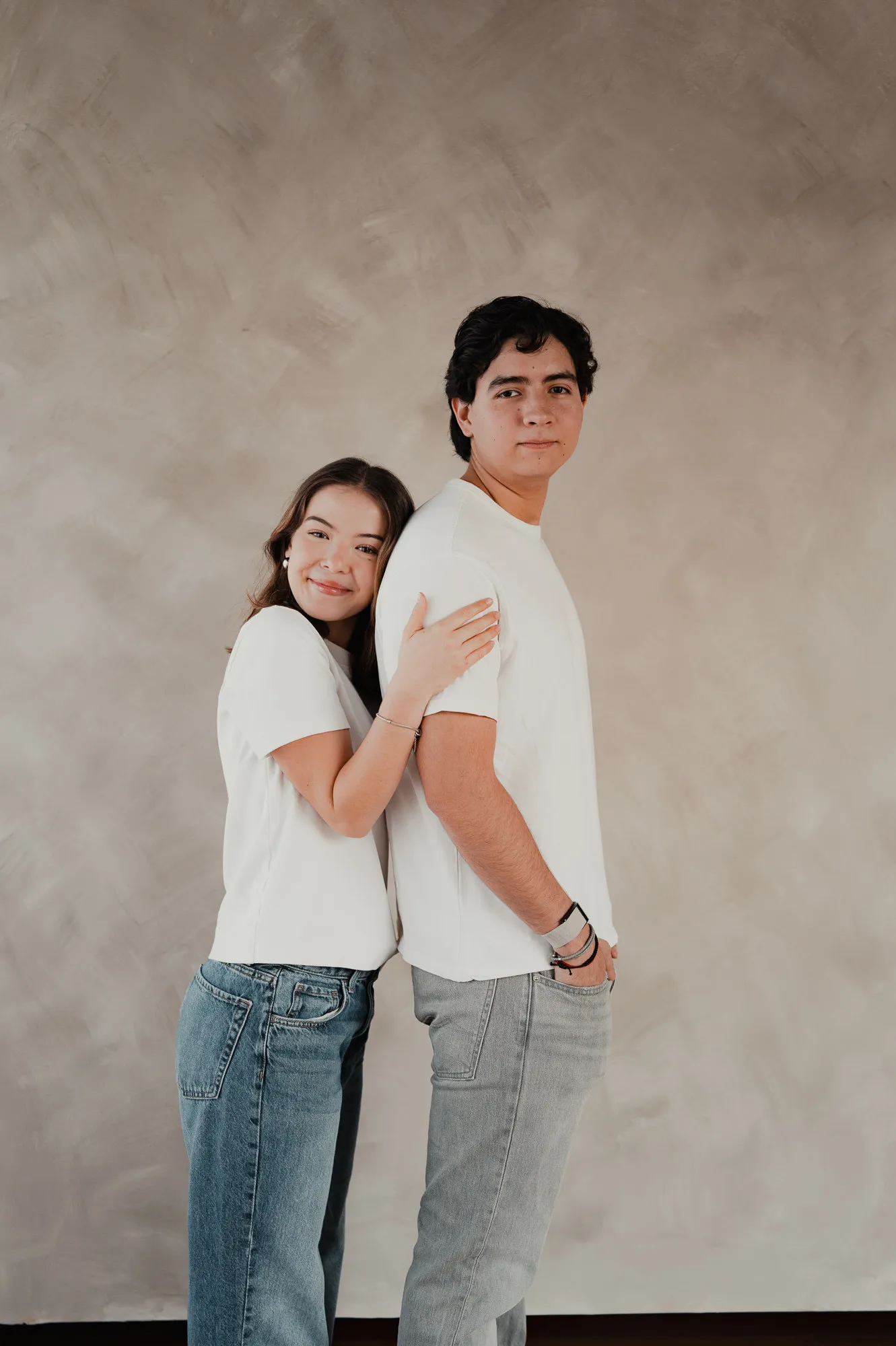 Una pareja posa abrazada con playera blanca y jeans frente a una pared color arena.