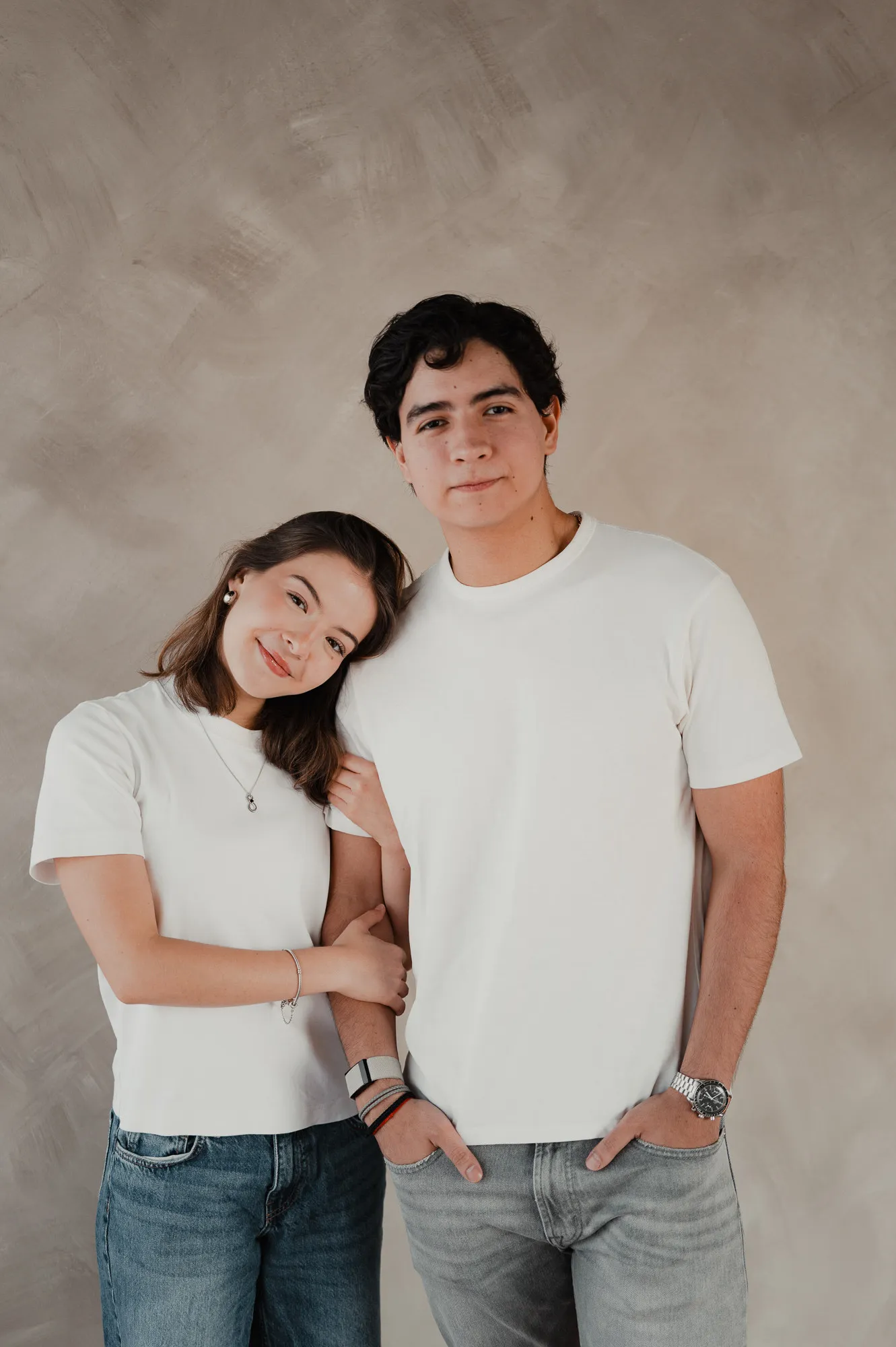 Una pareja posa abrazada con playera blanca y jeans frente a una pared color arena; ella inclina la cabeza.