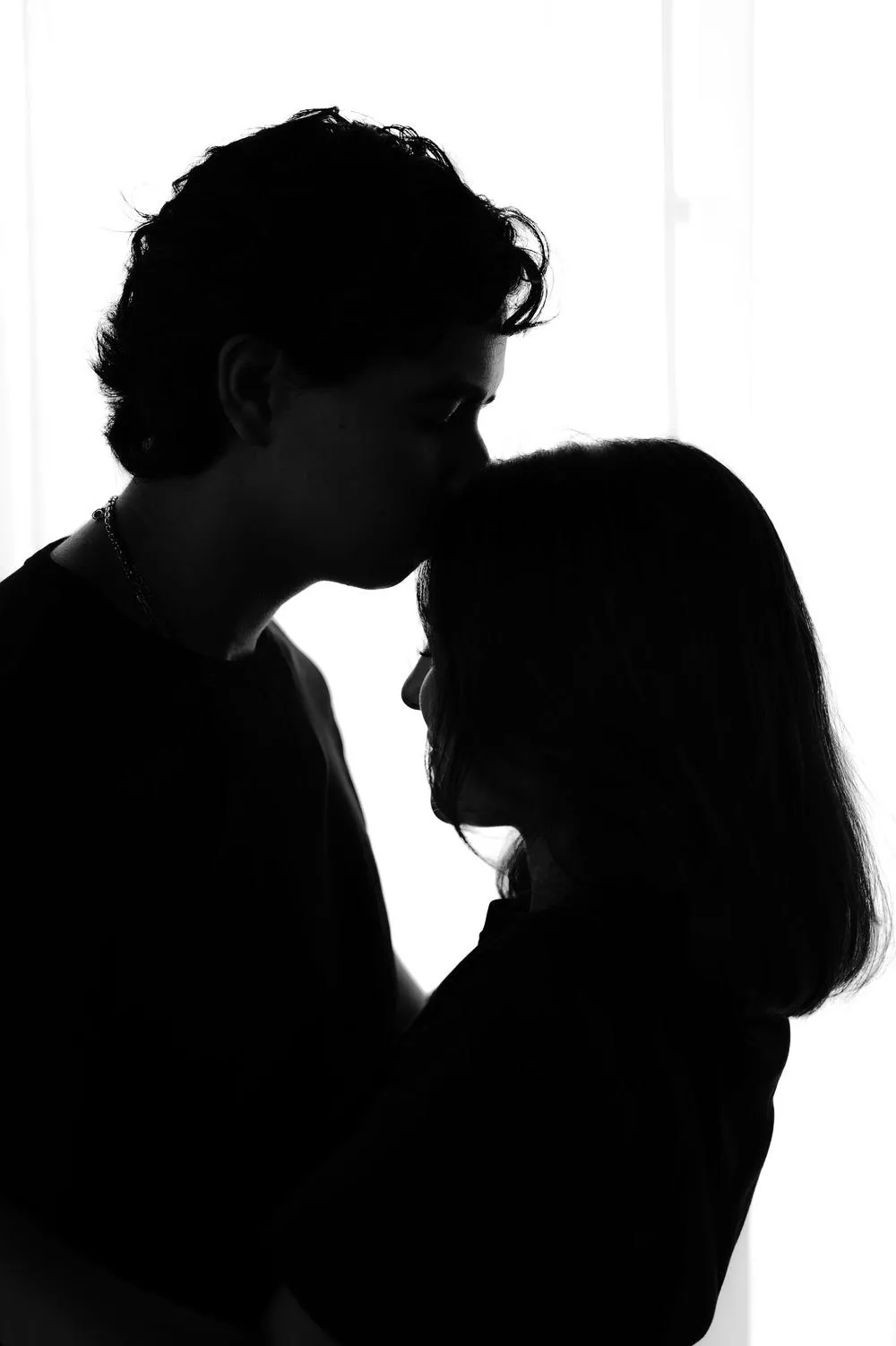En blanco y negro, la silueta de él besando la frente de ella, sobre fondo blanco.