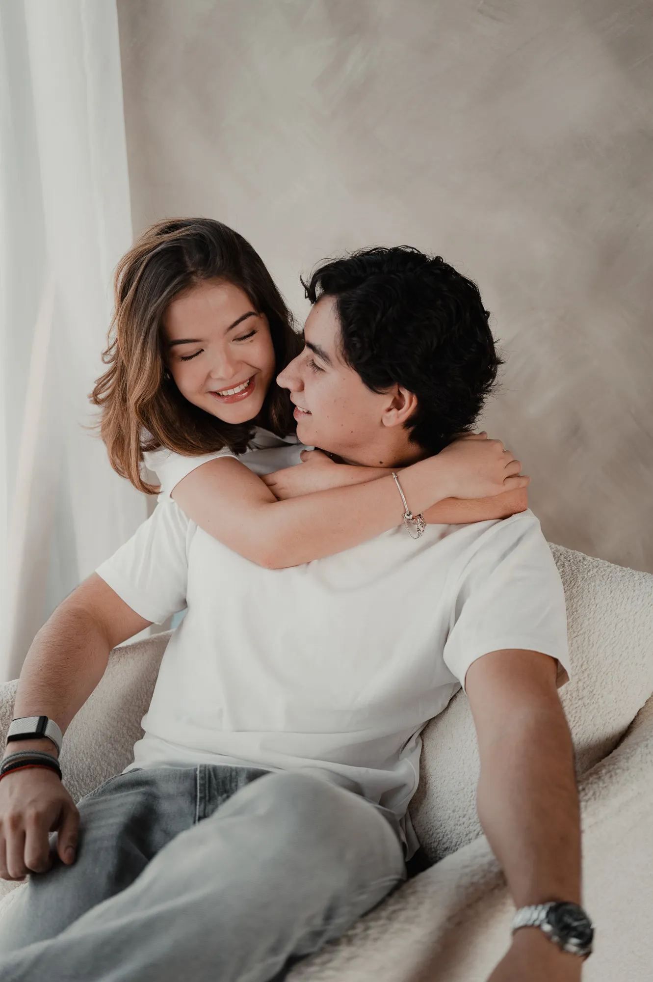 Ella abraza a su pareja por el cuello mientras ríen, sentados en un sillón claro.