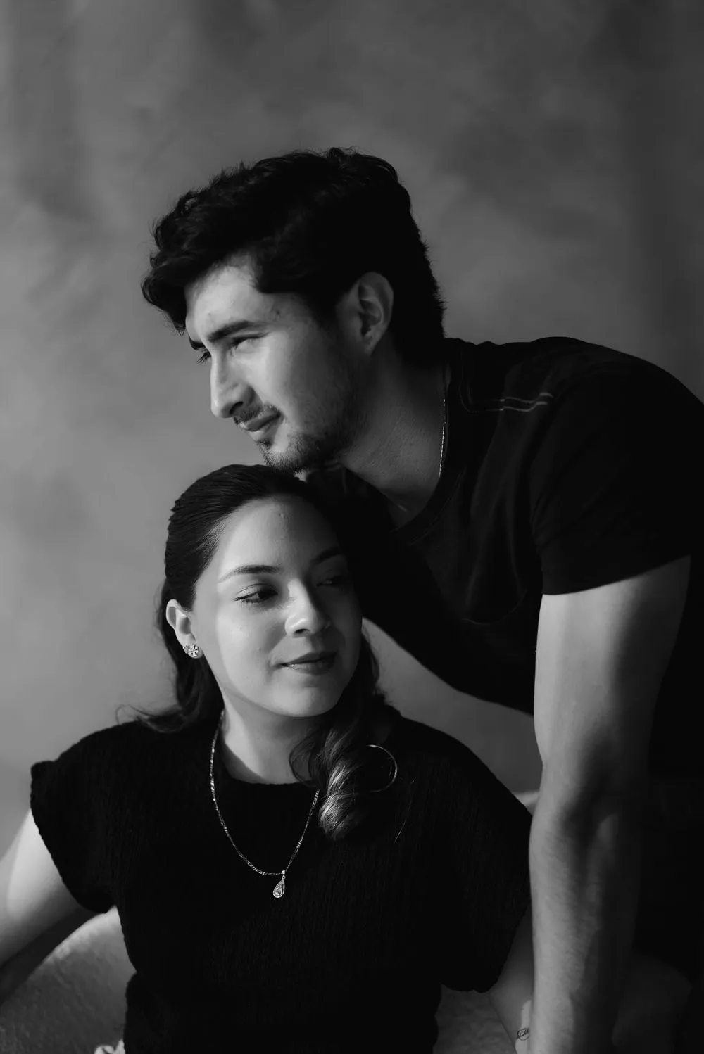 En blanco y negro, él inclina la cabeza sobre ella, que mira de frente, ambos con ropa oscura.