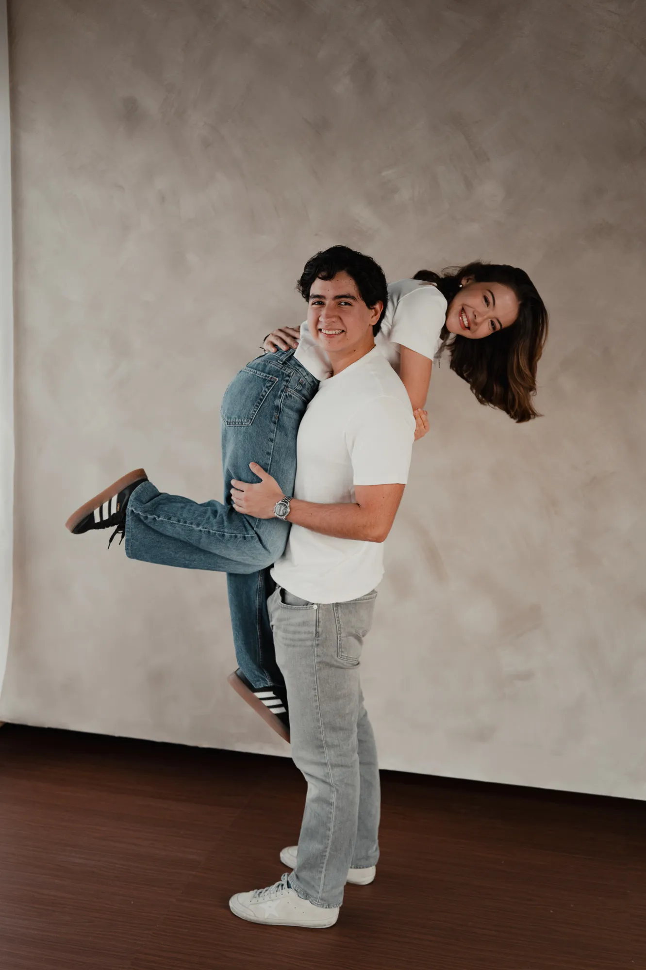 Él carga a su pareja en brazos mientras ella levanta las piernas, frente a una pared color arena.