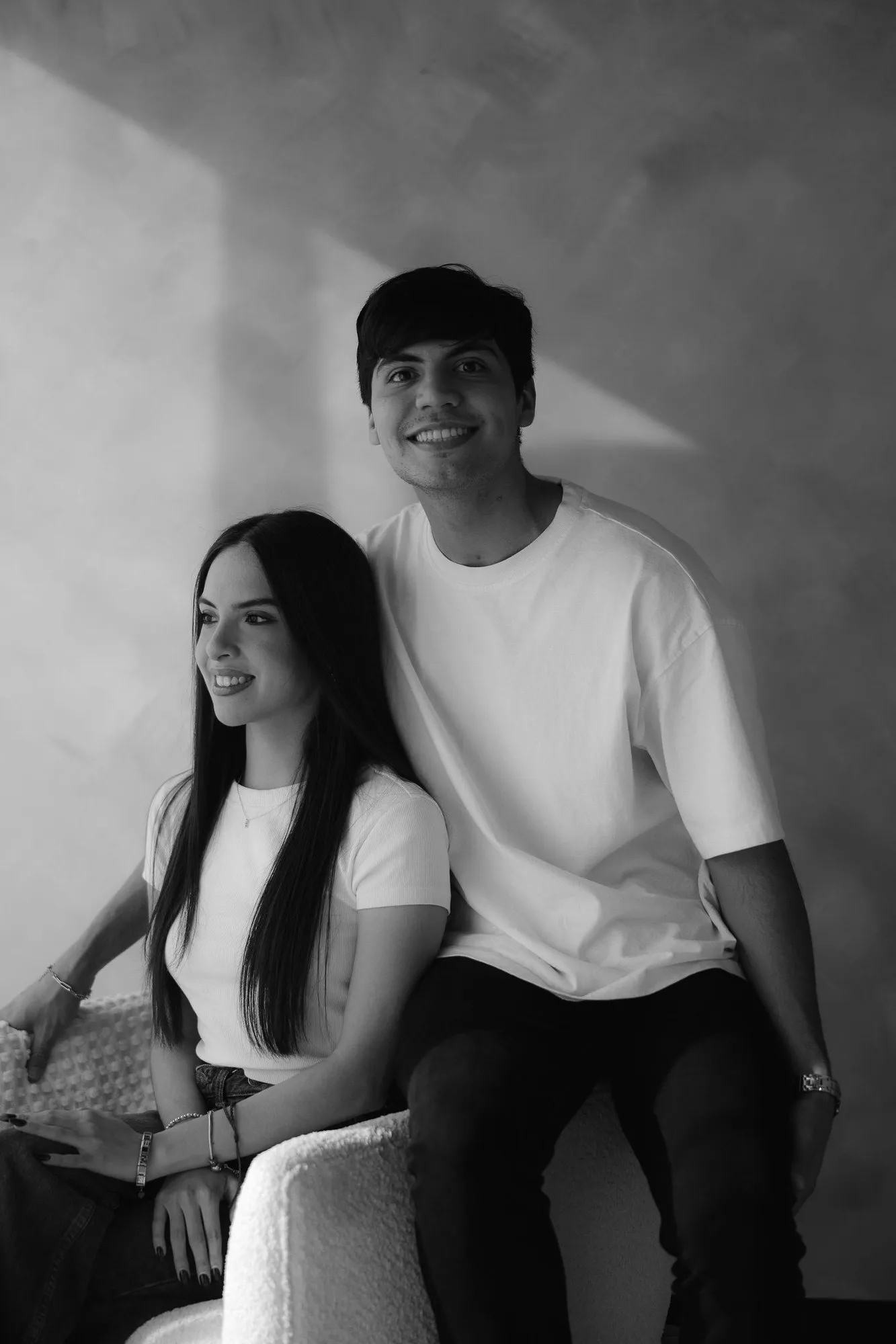 En blanco y negro, una pareja sentada se mira y sonríe, frente a una pared texturizada.